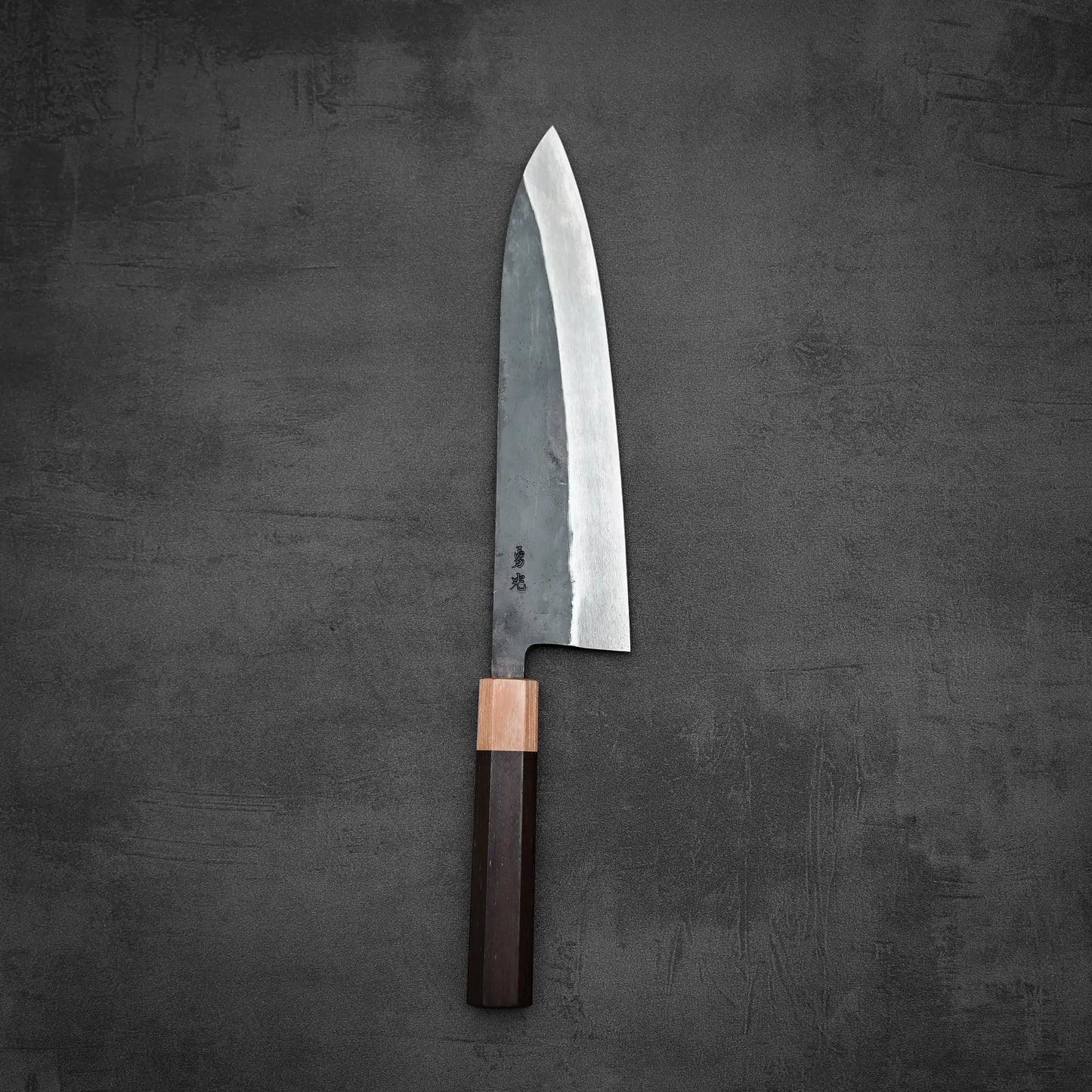 The Isamitsu kurouchi shirogami#1 gyuto 210mm, a large Japanese knife with a dark wooden handle and shiny blade, is displayed on a textured dark gray surface.
