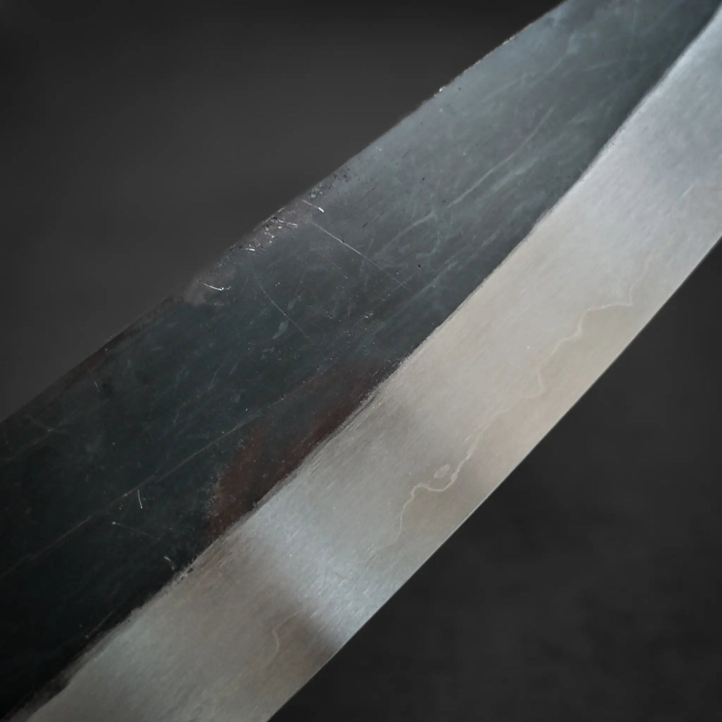 Close-up of the Isamitsu kurouchi shirogami#1 gyuto 210mm Japanese knife, highlighting the polished edge versus matte kurouchi finish. The dark, blurred background emphasizes Isamitsus unique blade texture and expert craftsmanship.