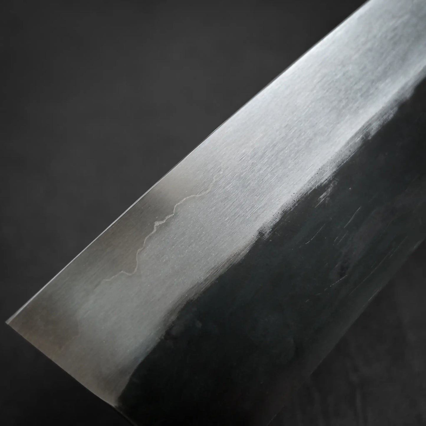 A close-up of the Isamitsu kurouchi shirogami#1 gyuto 210mm blade shows a clear transition from the polished edge to the dark spine, with subtle texture and a faint line revealing the metals patterning.