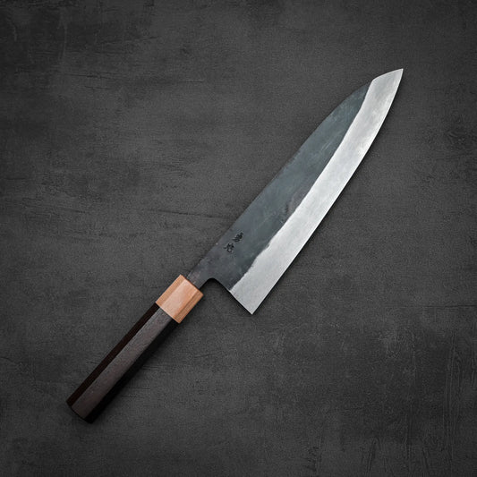 An Isamitsu kurouchi shirogami#1 gyuto 210mm Japanese chef’s knife by Isamitsu, featuring a dark wooden handle and large sharp blade, rests on a dark textured surface.
