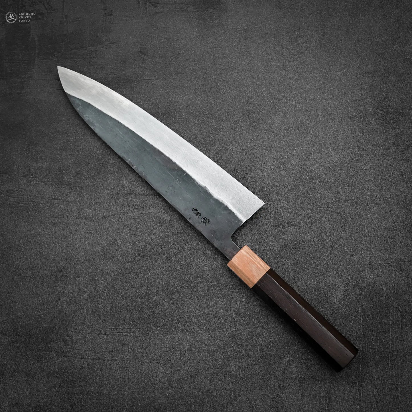 The Isamitsu kurouchi shirogami#1 gyuto 210mm chef’s knife by Isamitsu, featuring a dark wooden handle and silver blade, is placed diagonally from bottom right to top left on a dark gray textured surface.