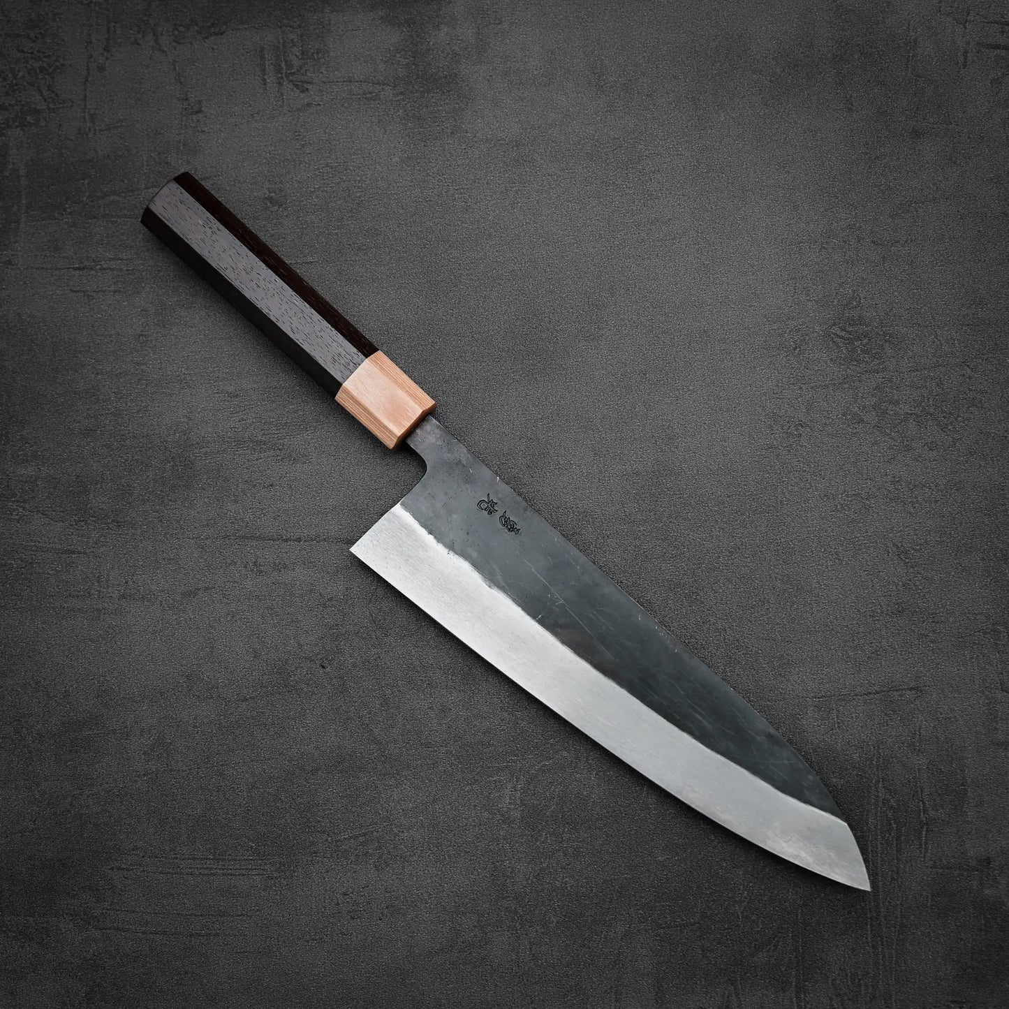 An Isamitsu kurouchi shirogami#1 gyuto 210mm by Isamitsu, featuring a dark wooden handle and wide, sharp blade, rests on a textured dark gray surface.