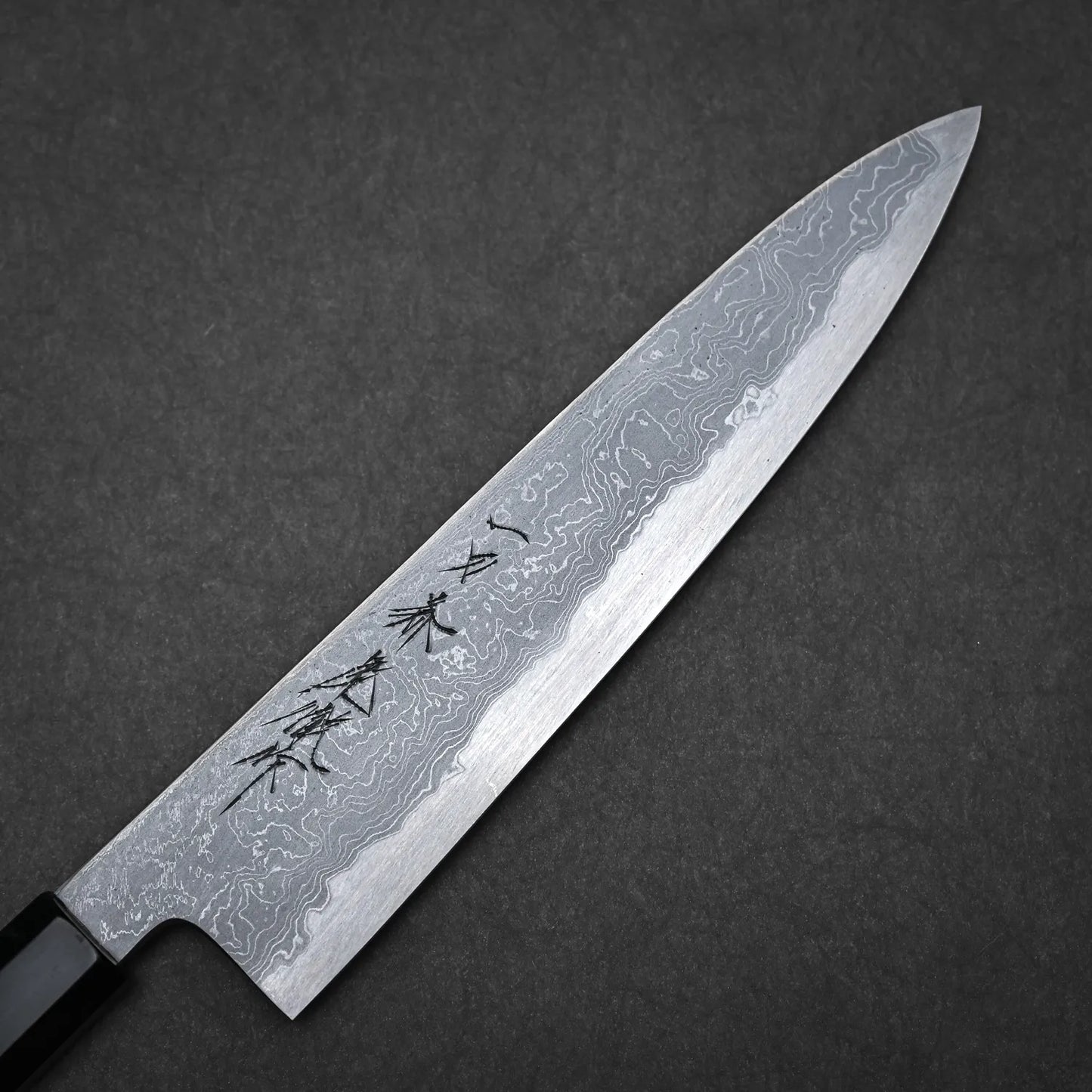 An Ittosai Kotetsu Damascus Aogami#1 Gyuto 240mm chef’s knife, displaying wavy patterns and engraved kanji, set against a dark textured background.