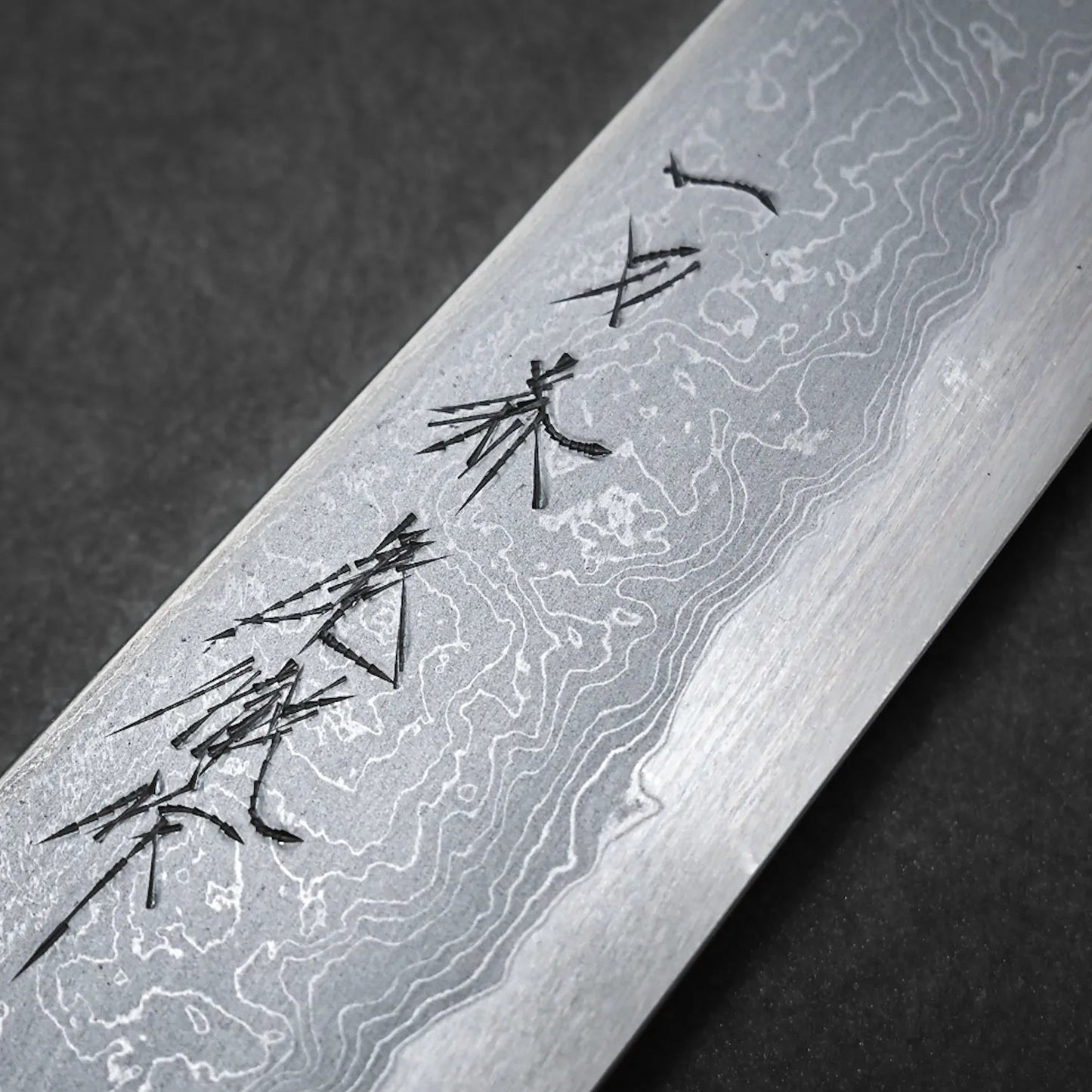 A close-up of the Ittosai Kotetsu damascus aogami#1 gyuto 240mm shows detailed layered patterns and engraved kanji on its blade, handcrafted in Sakai, Japan, set against a dark background.