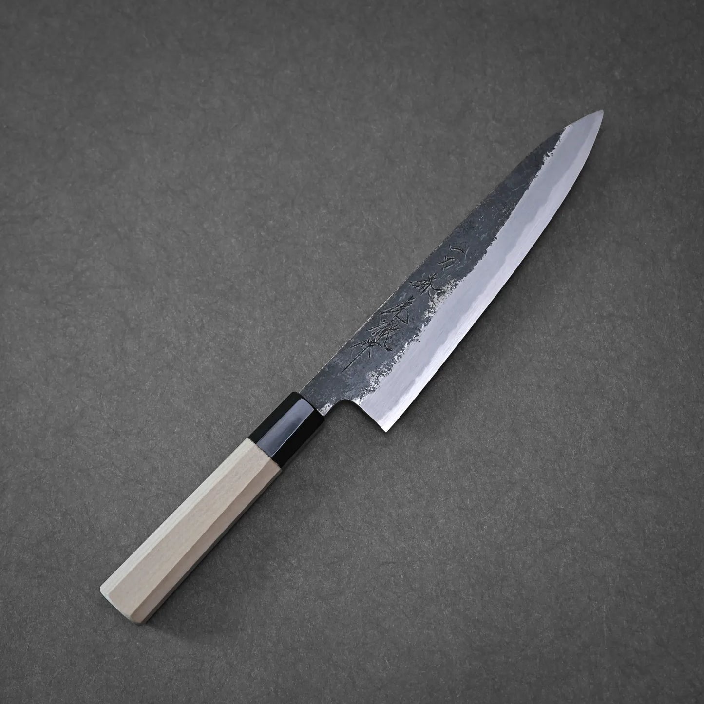 The Yoshikazu Tanaka Ittosai Kotetsu Gokujo kurouchi shirogami#1 gyuto 210mm, featuring a light wooden handle and dark blade, rests on a gray surface.