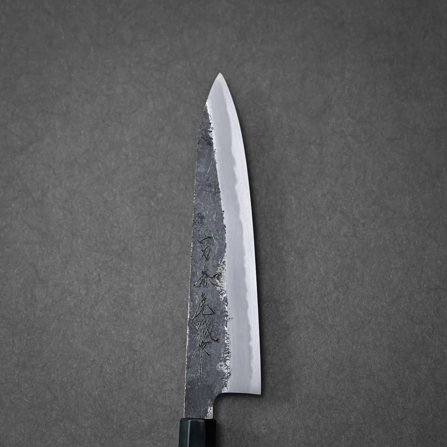 The Yoshikazu Tanaka Ittosai Kotetsu Gokujo kurouchi shirogami#1 gyuto 210mm chefs knife, with a sharp, pointed blade and hammered finish, features a polished edge and engraved markings on dark grey.