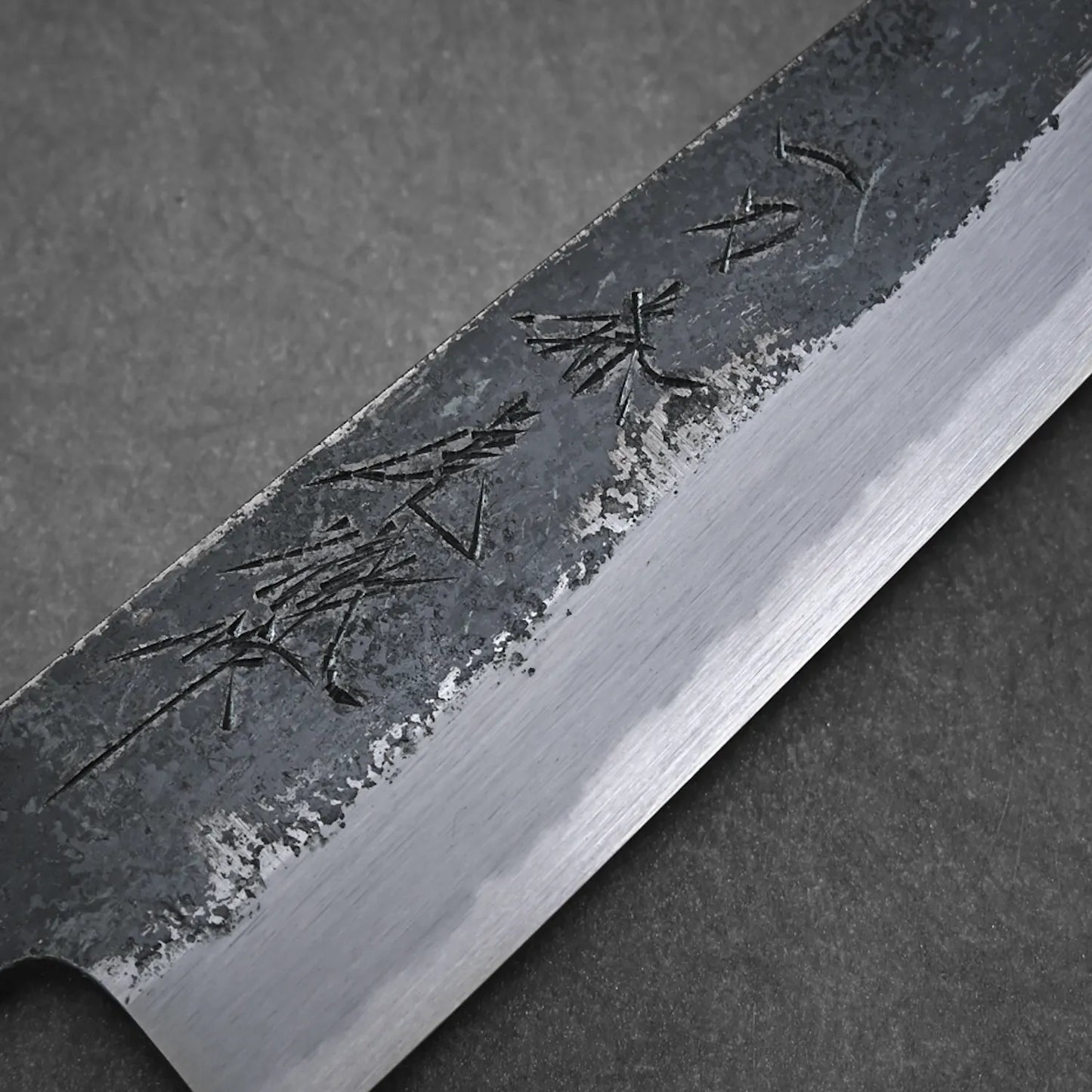A close-up of the Yoshikazu Tanaka Ittosai Kotetsu Gokujo kurouchi shirogami#1 gyuto 210mm shows a partly rough kurouchi finish, polished edge, and Japanese characters on the blade, set against a gray textured background.