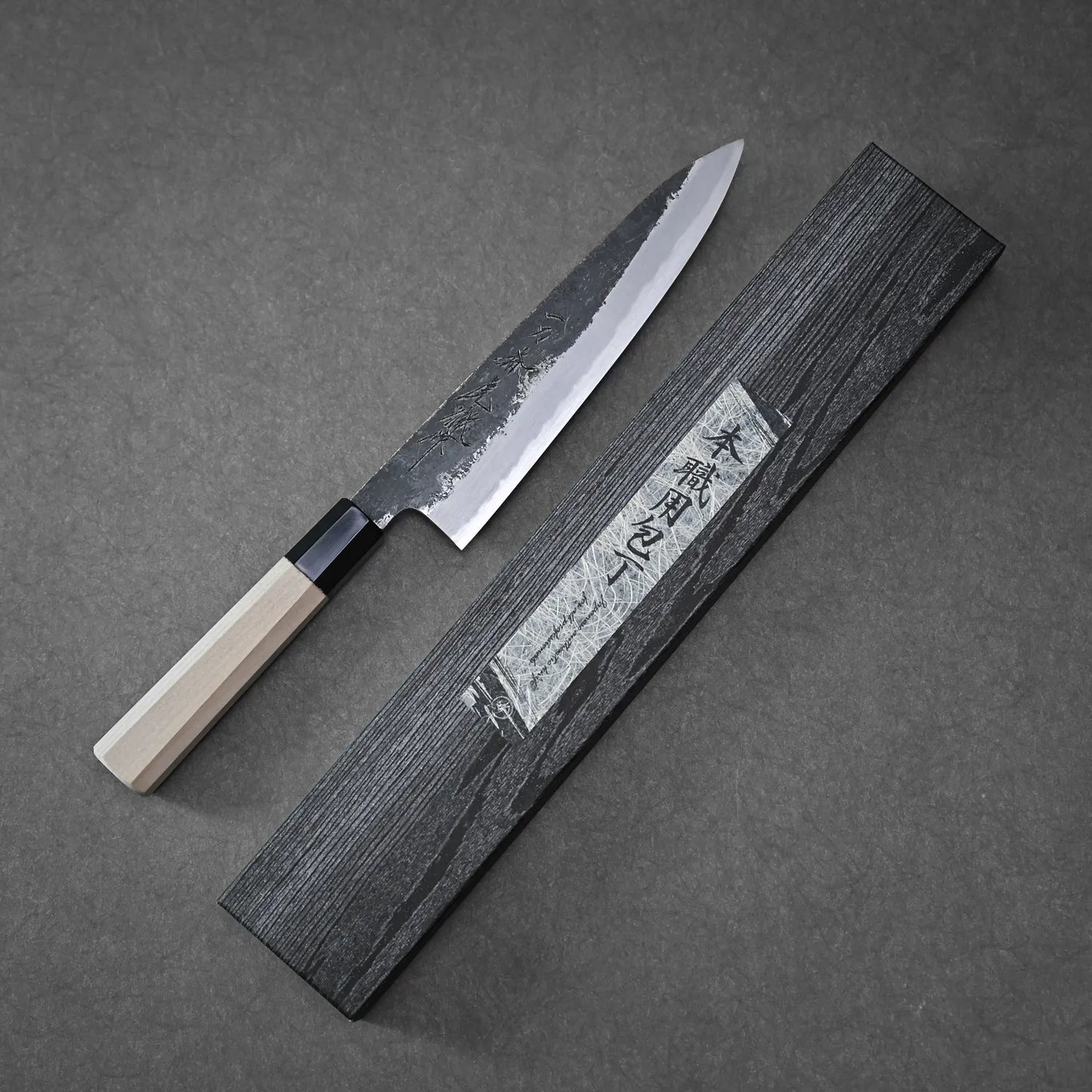 The Yoshikazu Tanaka Ittosai Kotetsu Gokujo kurouchi shirogami#1 gyuto 210mm, with a light wooden handle, rests beside a dark gray knife box with Japanese writing on the lid, displayed on a gray surface.