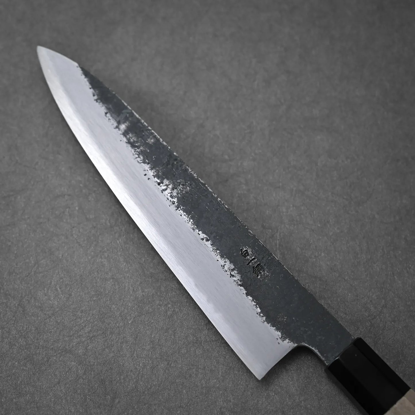 The Yoshikazu Tanaka Ittosai Kotetsu Gokujo kurouchi shirogami#1 gyuto 210mm, showcasing a pointed tip and striking finish transition, is displayed on a gray surface with its dark kurouchi upper blade and polished lower edge.