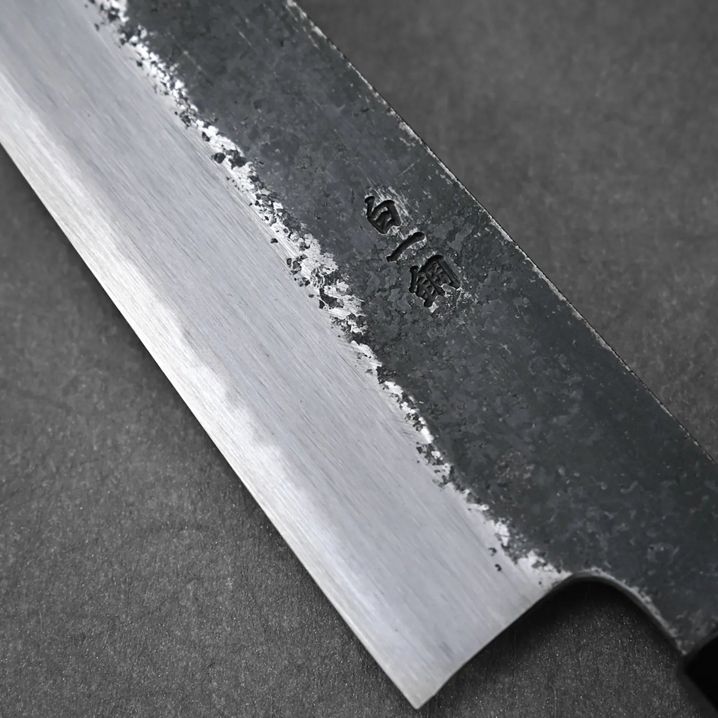Close-up of the Ittosai Kotetsu Gokujo kurouchi shirogami#1 gyuto 210mm by Yoshikazu Tanaka, showing a sharp, polished edge and kurouchi finish with engraved kanji characters. The blade rests on a dark, matte background.
