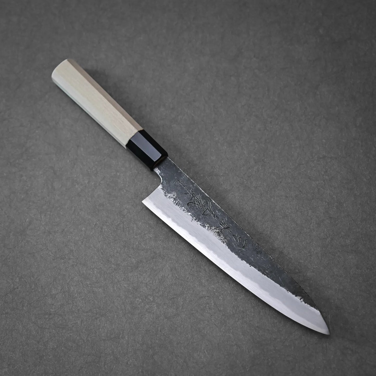 The Yoshikazu Tanaka Ittosai Kotetsu Gokujo Kurouchi Shirogami #1 Gyuto 210mm, featuring a light wooden handle and dark kurouchi finish blade, is placed on a gray surface.