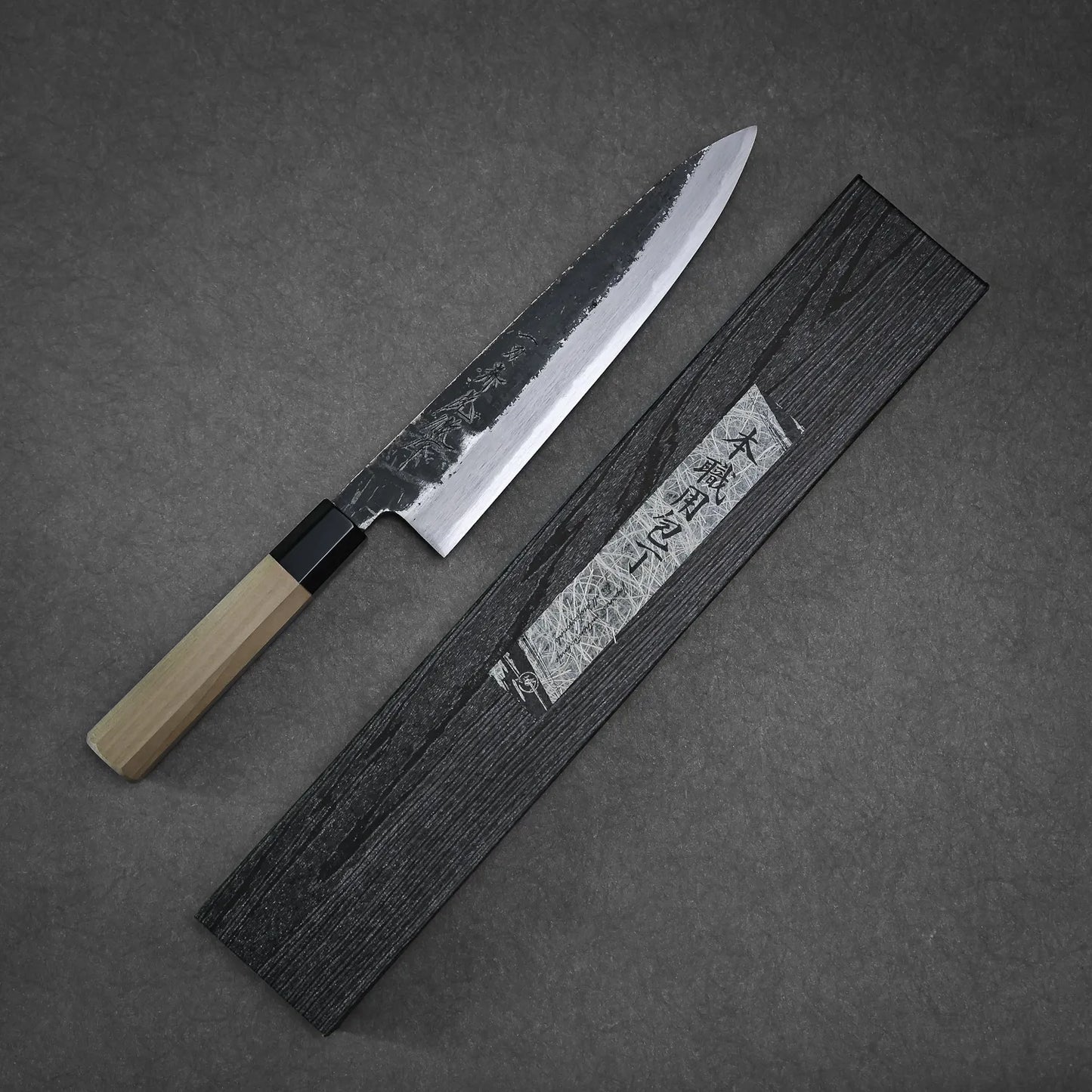 A Yoshikazu Tanaka Ittosai Kotetsu Gokujo kurouchi shirogami#1 gyuto 240mm chefs knife with a wooden handle lies diagonally next to a black box with Japanese characters, set against a gray background.