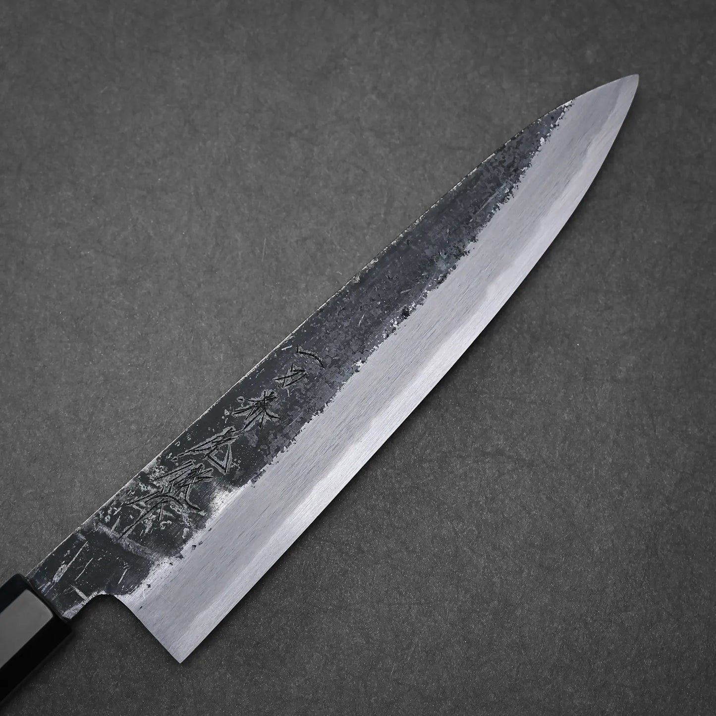 The Yoshikazu Tanaka Ittosai Kotetsu Gokujo kurouchi shirogami#1 gyuto 240mm, featuring a polished Shirogami #1 edge and textured, engraved upper blade, rests on a dark surface.