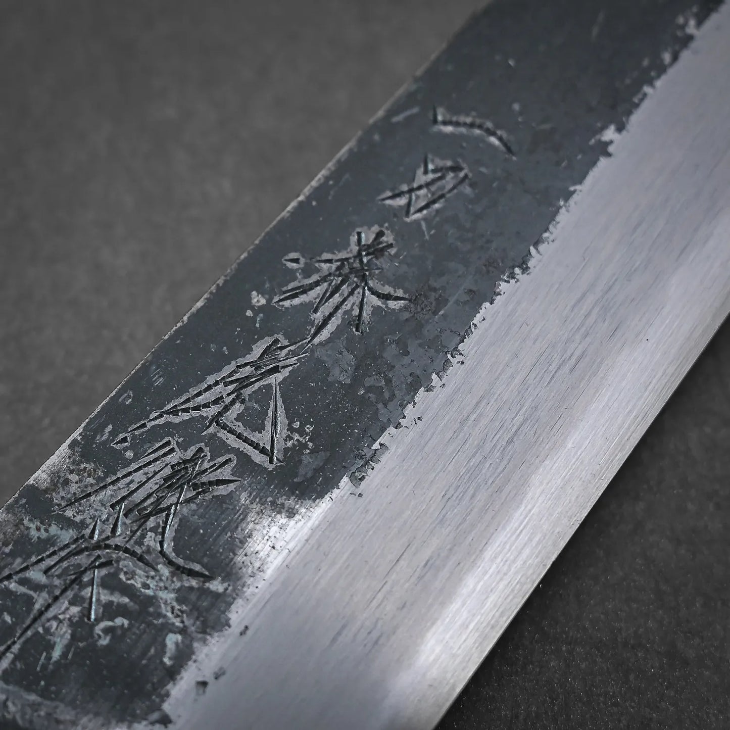 Close-up of the Yoshikazu Tanaka Ittosai Kotetsu Gokujo kurouchi shirogami#1 gyuto 240mm, showing its kurouchi finish, Shirogami #1 core steel, and Japanese characters near the textured blade contrasting with the polished edge.
