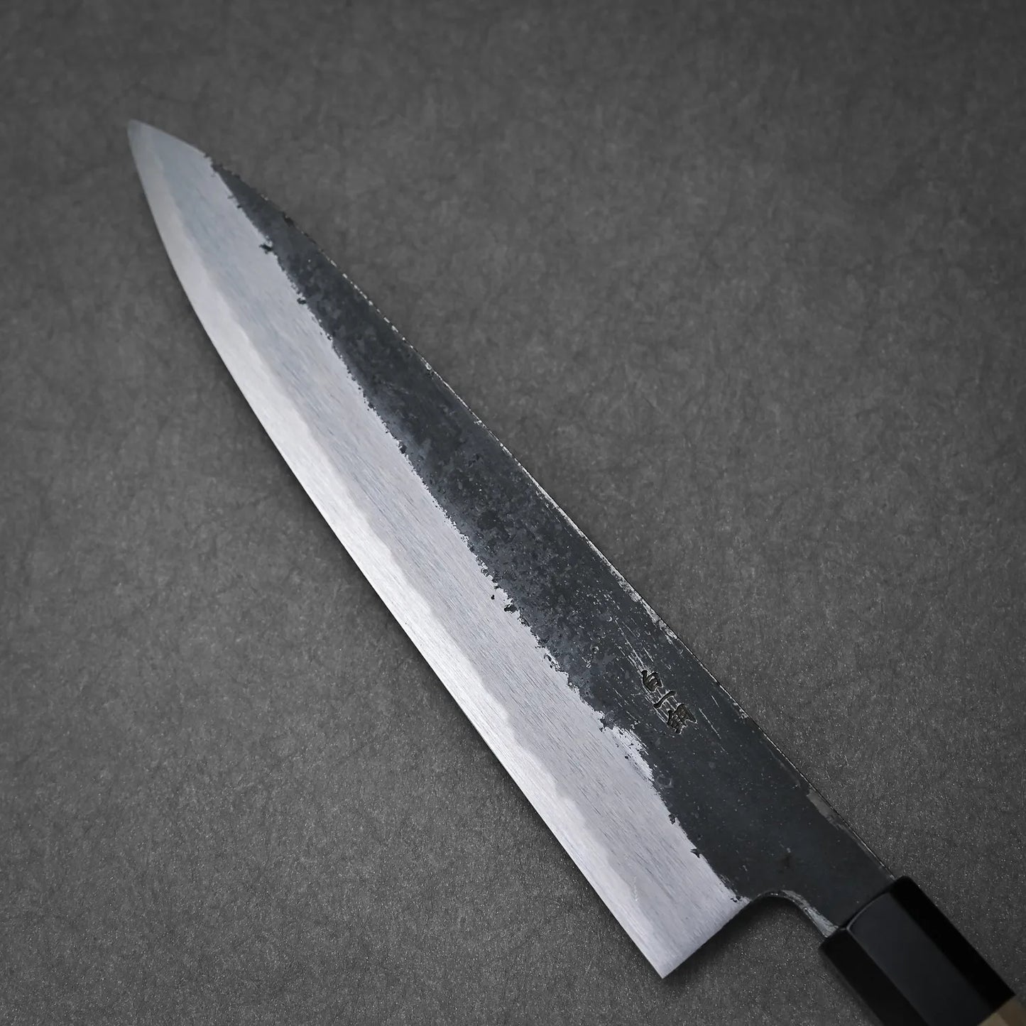 A close-up of the Yoshikazu Tanaka Ittosai Kotetsu Gokujo kurouchi shirogami#1 gyuto 240mm shows its sharp blade with a Shirogami #1 core and textured upper section, resting on a grey surface.