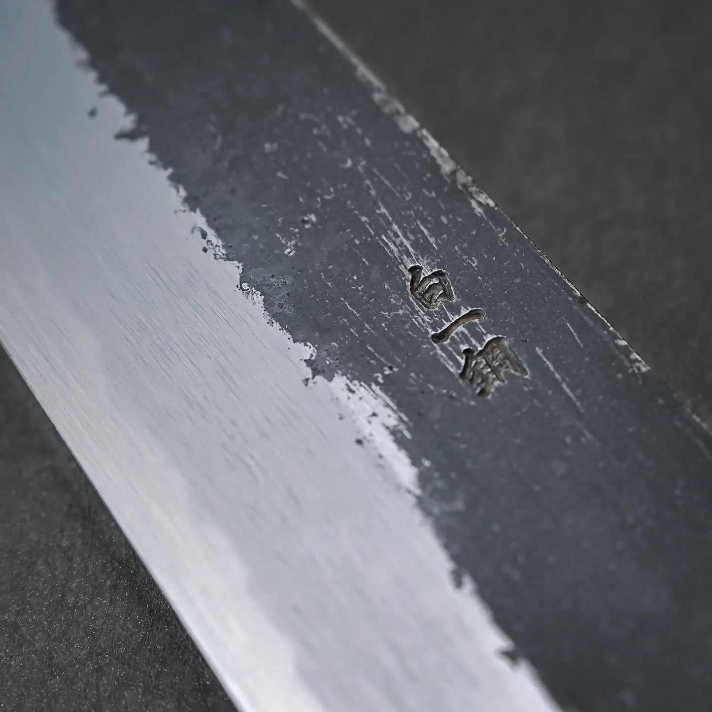 Close-up of a Yoshikazu Tanaka Ittosai Kotetsu Gokujo kurouchi shirogami#1 gyuto 240mm blade, showing textured steel and engraved characters on the Shirogami #1 core against a dark, blurred background.