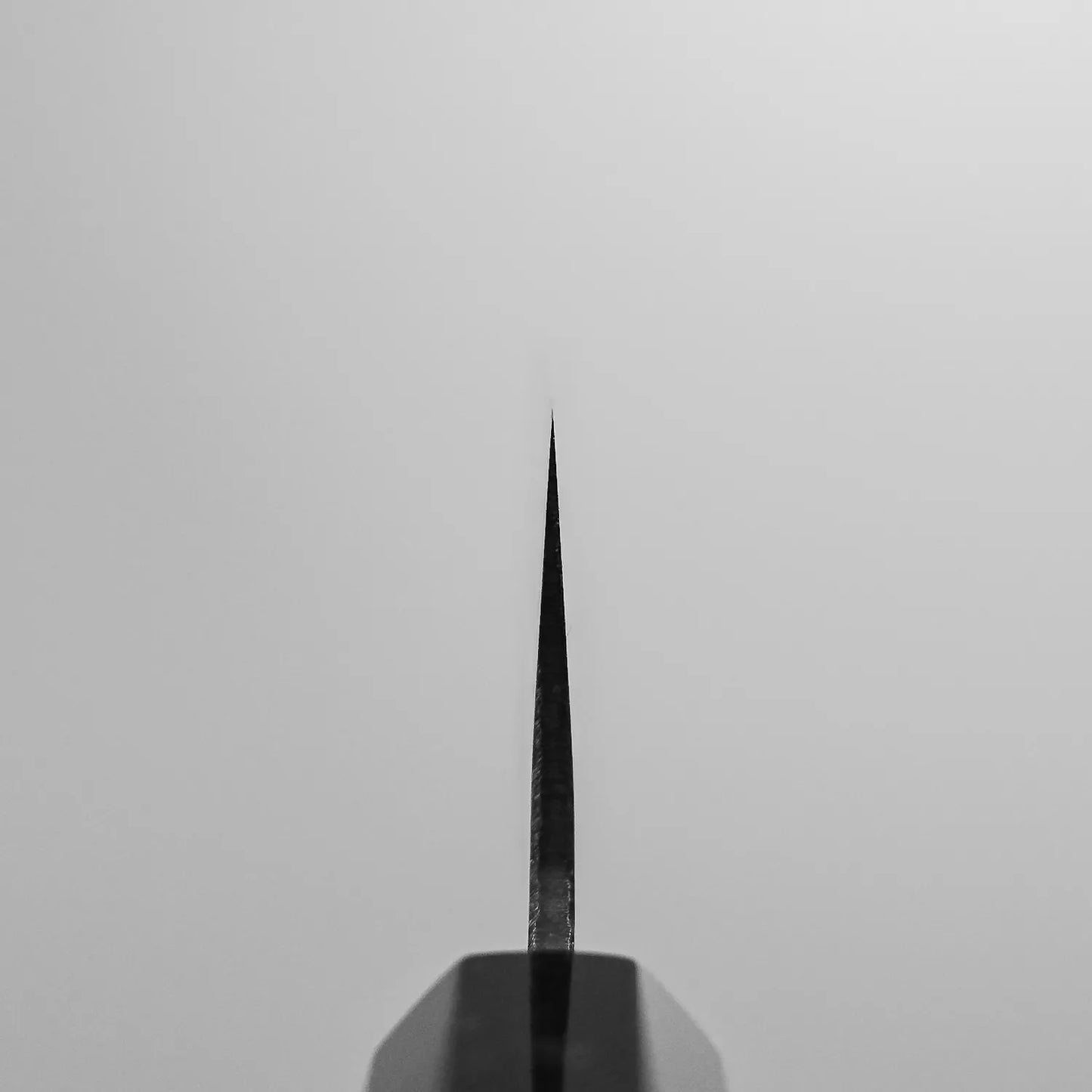 A centered close-up shows the tip of the Yoshikazu Tanaka Ittosai Kotetsu Gokujo kurouchi shirogami#1 gyuto 240mm chef’s knife, its sharp edge vertical against a plain light gray background, with part of the handle visible at the bottom.