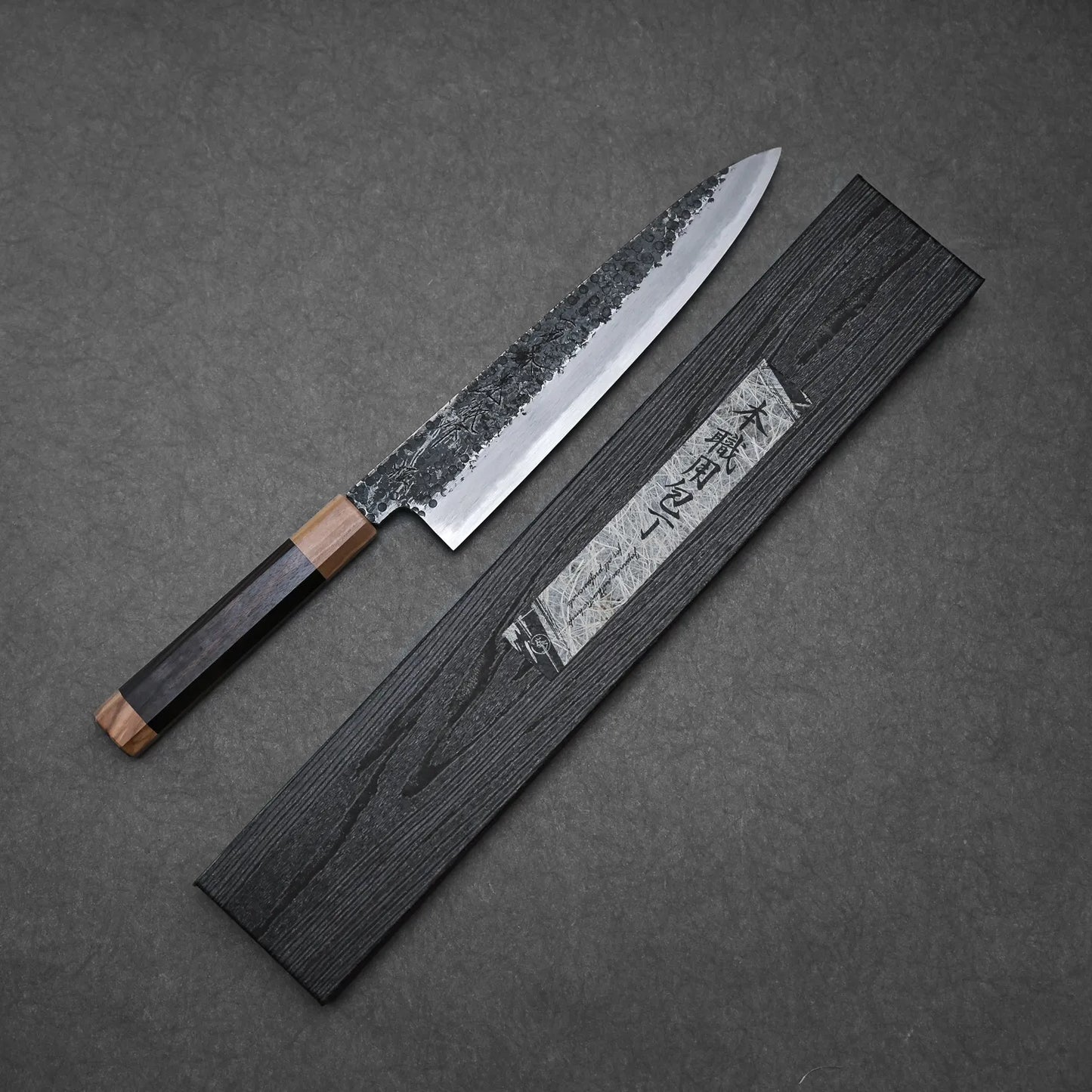 A Yoshikazu Tanaka Ittosai Kotetsu tsuchime kurouchi shirogami#1 gyuto 240mm with a wooden handle and hammered blade rests beside a black sheath with Japanese characters, all displayed on a dark gray surface.