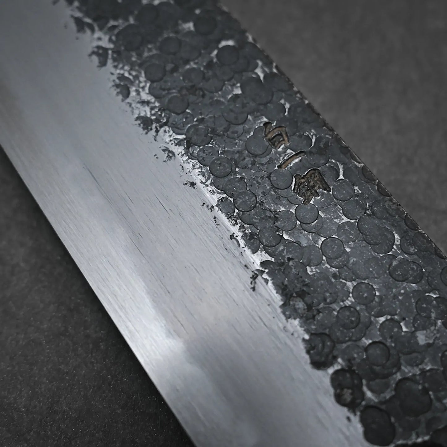Close-up of the Yoshikazu Tanaka Ittosai Kotetsu tsuchime kurouchi shirogami#1 gyuto 240mm, forged in Sakai, Japan, showing its hammered black upper blade and smooth polished edge with engraved Japanese characters.
