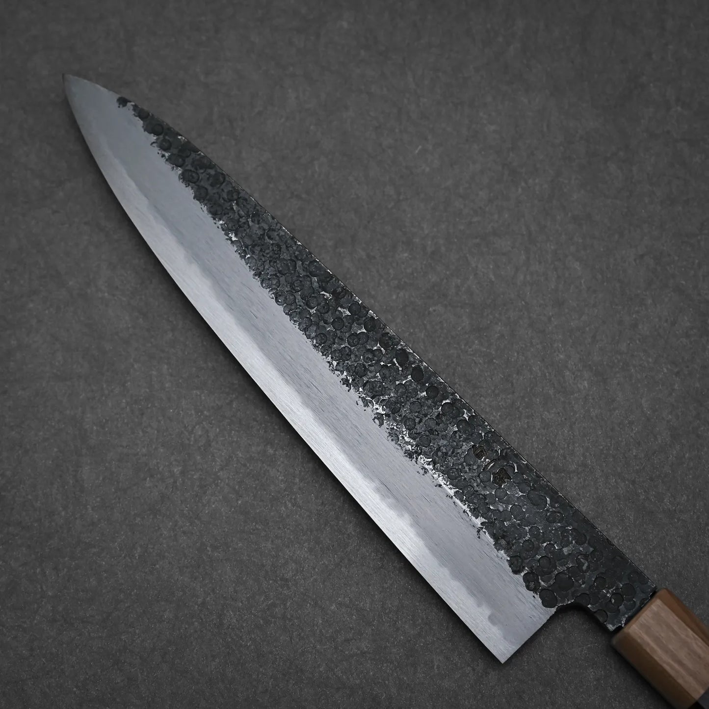 The Yoshikazu Tanaka Ittosai Kotetsu tsuchime kurouchi shirogami#1 gyuto 240mm chef’s knife features a hammered blade and sharp Shirogami #1 steel edge, shown against a dark gray background.
