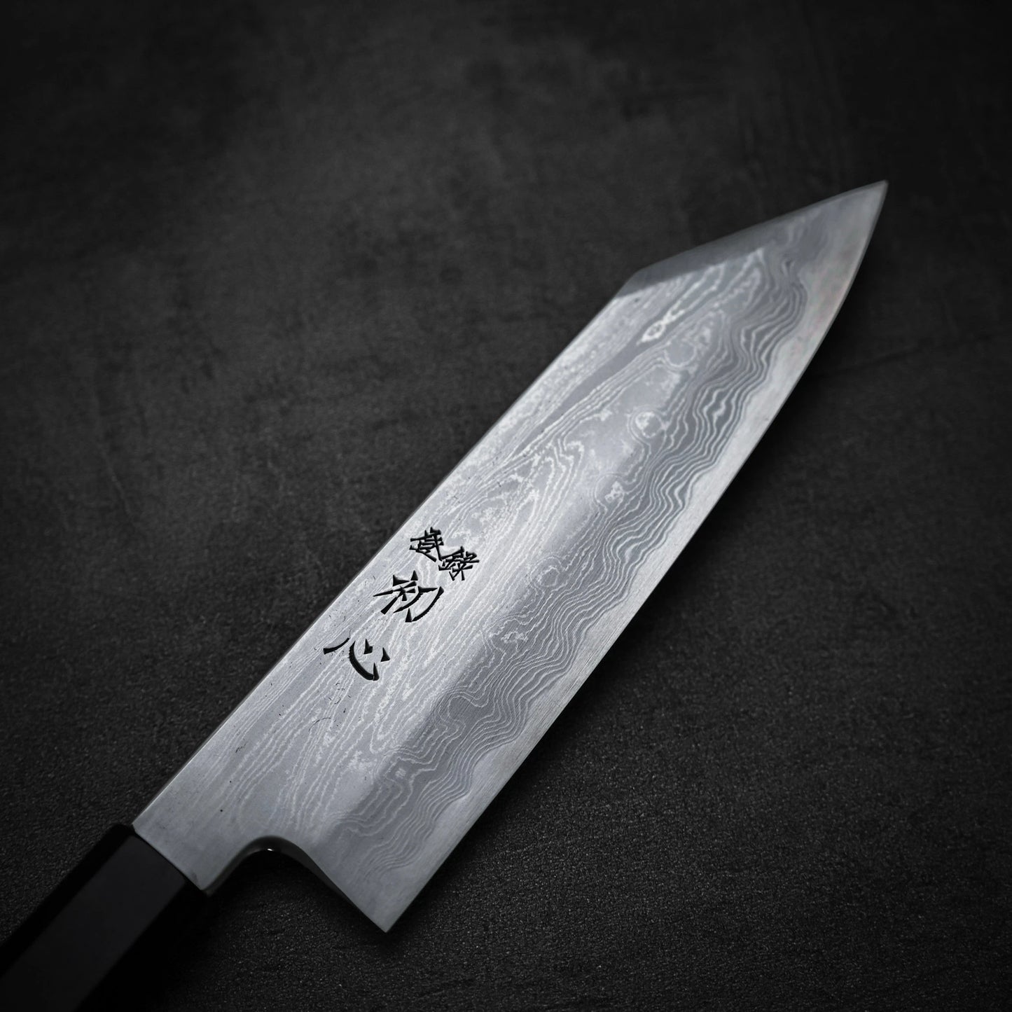 A close-up of the Satoshi Nakagawa Nakagawa aogami#1 damascus bunka 180mm, featuring a Damascus steel blade with wave patterns and Japanese characters near the handle, displayed on a dark textured surface.
