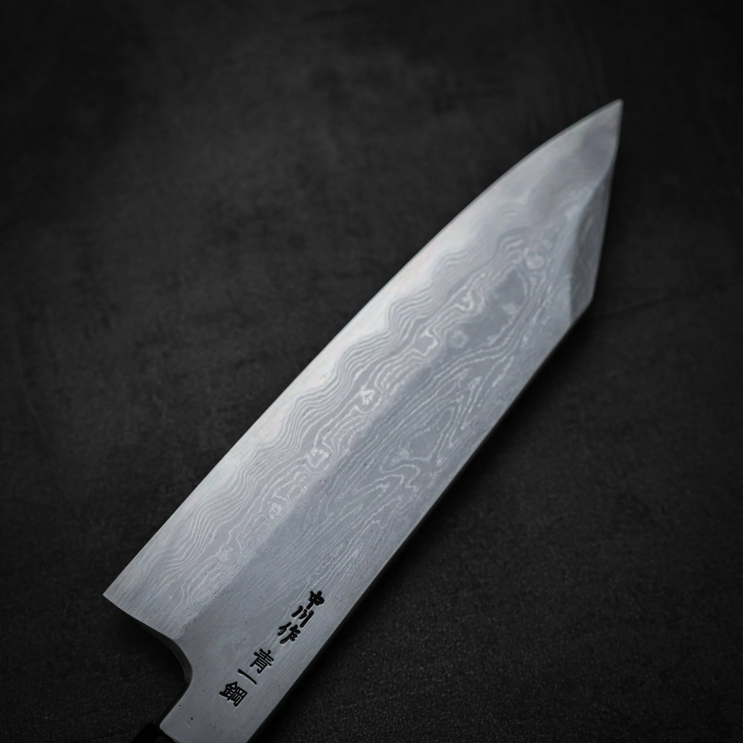 A close-up of the Satoshi Nakagawa Nakagawa aogami#1 damascus bunka 180mm knife, featuring intricate Damascus steel patterns on the blade and engraved Japanese characters near the handle, set against a dark textured background.
