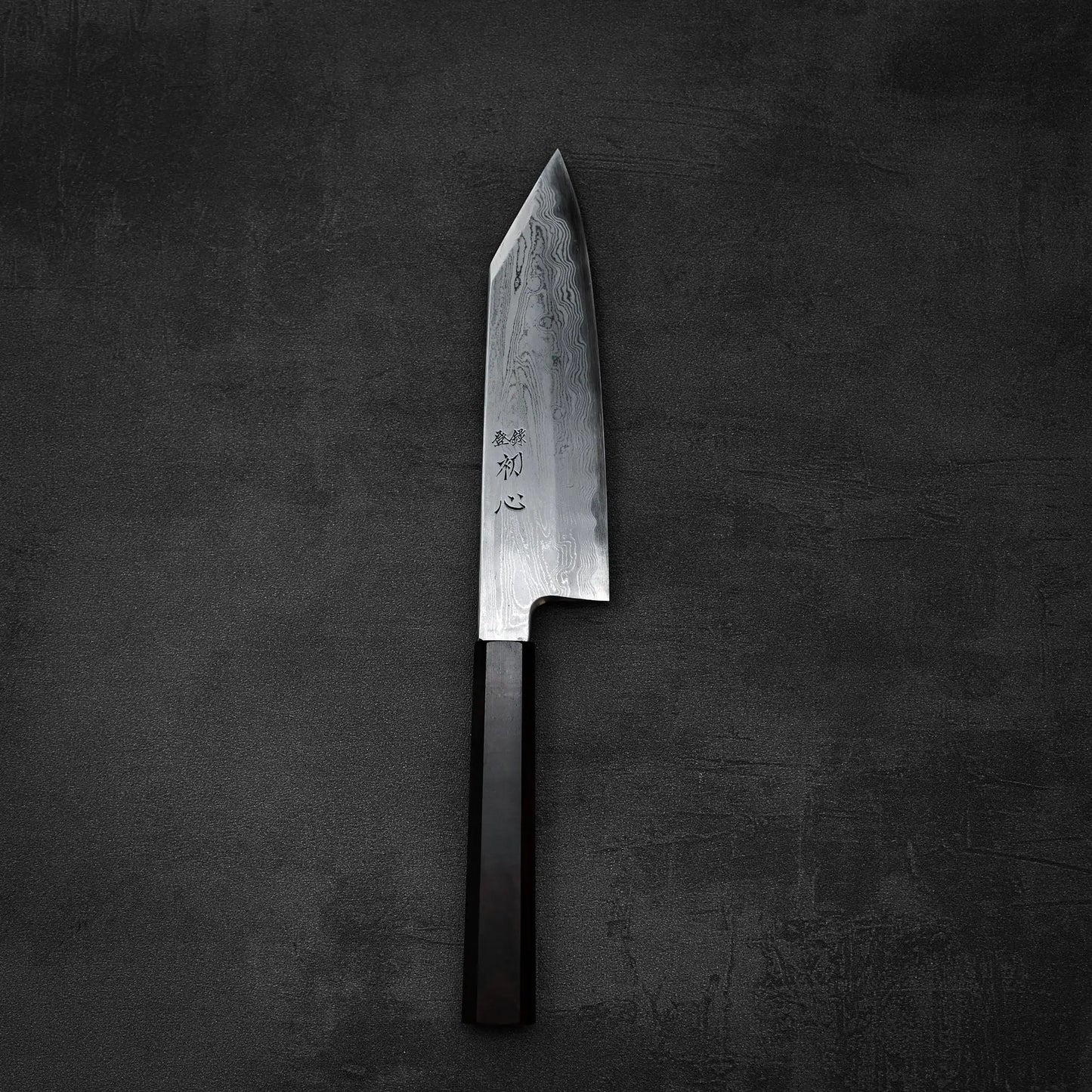 The Satoshi Nakagawa aogami#1 damascus bunka 180mm features a patterned steel blade with Japanese characters near its dark handle, and rests on a textured dark gray surface.
