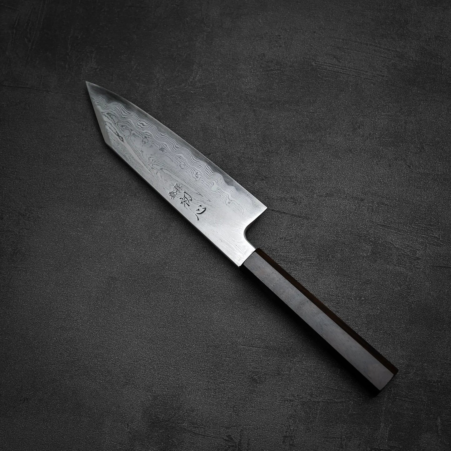 The Satoshi Nakagawa aogami#1 damascus bunka 180mm, featuring a pointed blade with elegant patterns and engraved Japanese characters and a wooden handle, is placed diagonally on a dark, textured surface.