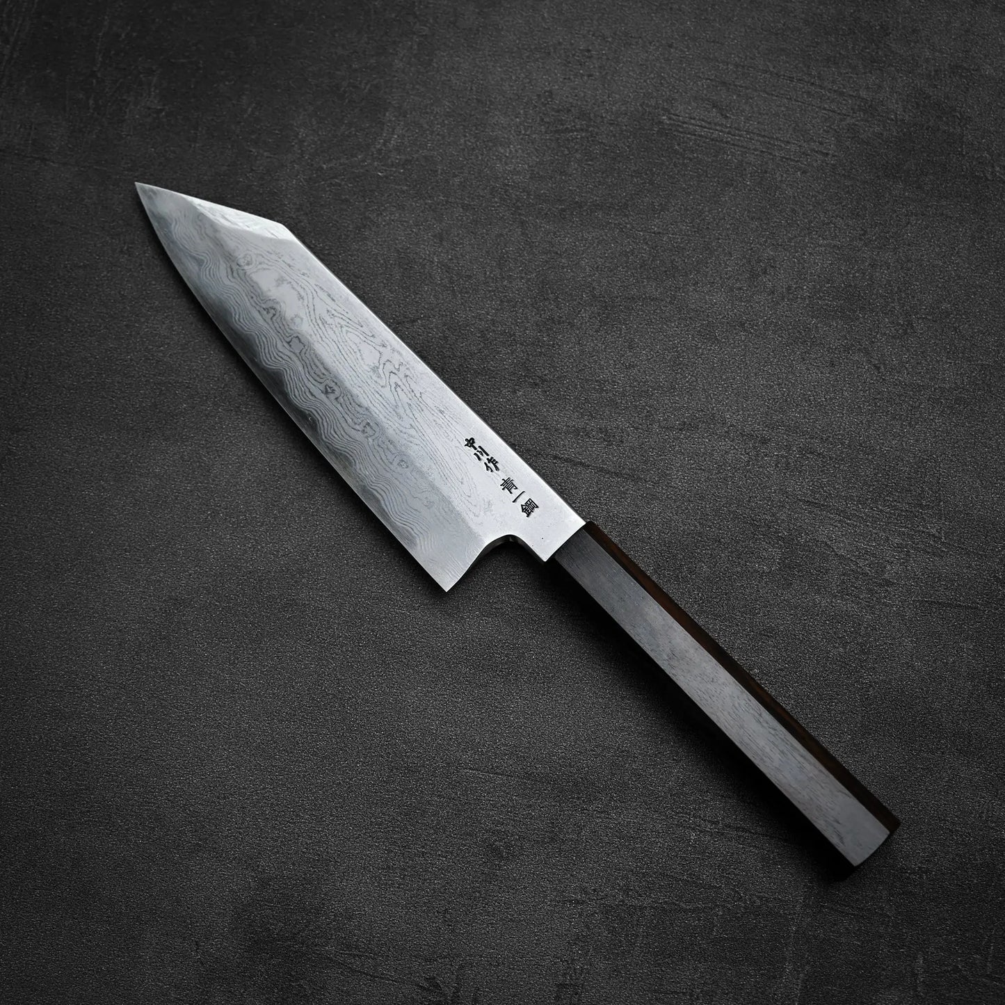 The Satoshi Nakagawa Nakagawa aogami#1 damascus bunka 180mm, featuring a damascus-patterned blade and dark wooden handle, rests on a dark textured surface.