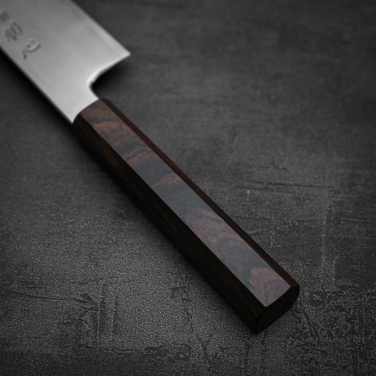 Close-up of the Satoshi Nakagawa Nakagawa aogami#1 damascus bunka 180mm knife, featuring its polished dark wooden handle resting on a dark textured surface. Part of the blade and the full handle are visible.