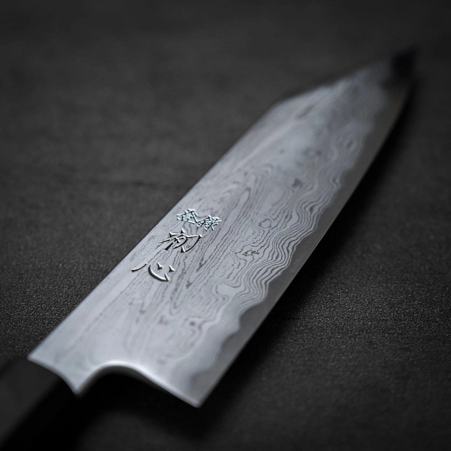 A close-up of the Satoshi Nakagawa Nakagawa aogami#1 damascus bunka 180mm highlights its Damascus steel patterns and engraved kanji on the blade, resting on a dark, textured surface.