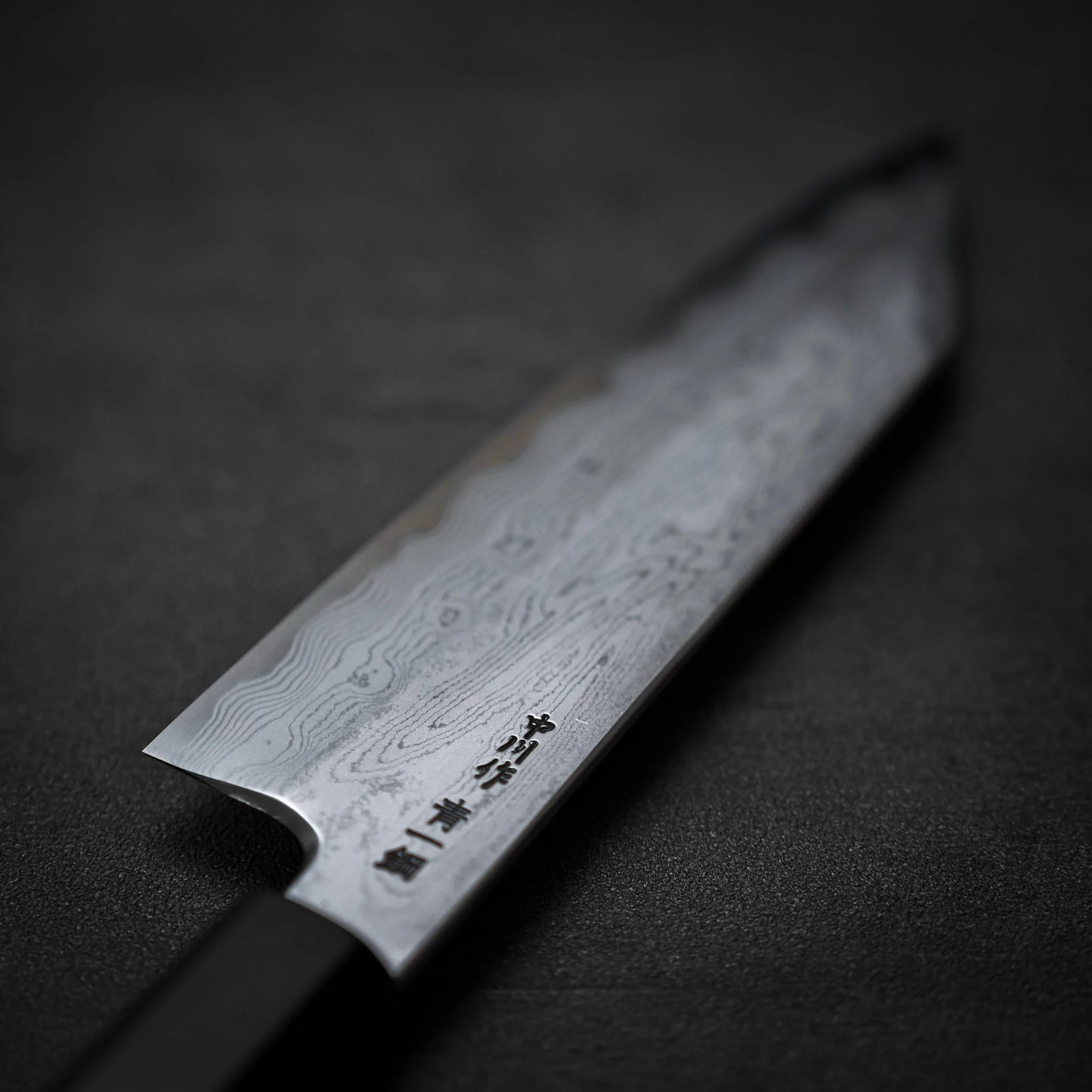 A close-up of the Satoshi Nakagawa aogami#1 damascus bunka 180mm knife shows its wavy Damascus steel blade and engraved Japanese characters, set against a dark, blurred background.