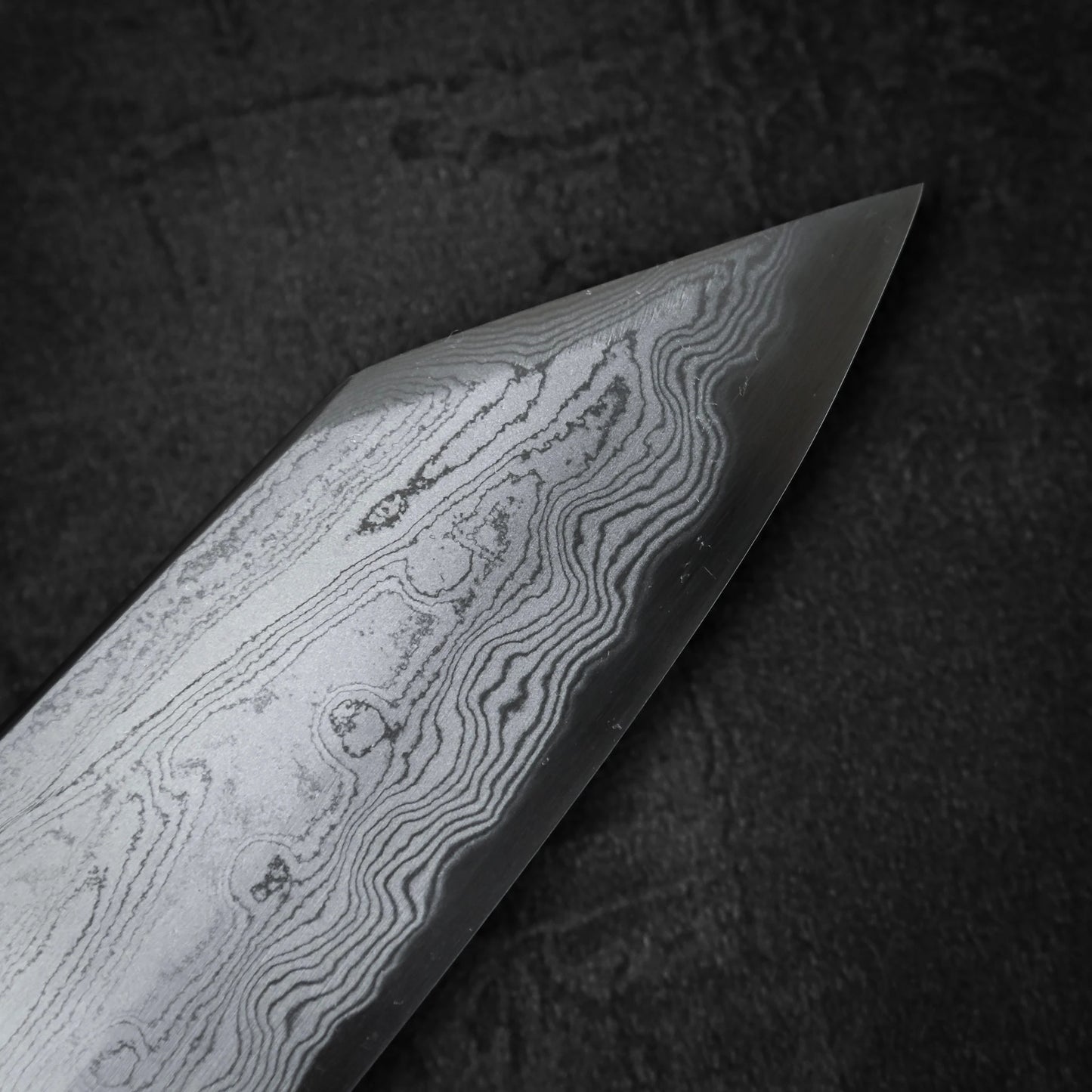 A close-up of the Satoshi Nakagawa aogami#1 damascus bunka 180mm reveals striking wavy layers, emphasizing the craftsmanship of this Japanese knife set against a dark, textured backdrop.