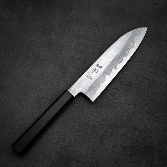 The Satoshi Nakagawa Nakagawa aogami#1 damascus santoku 180mm, featuring a wavy damascus finish and Japanese characters near the handle, rests on a black textured surface. Its dark rectangular handle highlights its Japanese design.