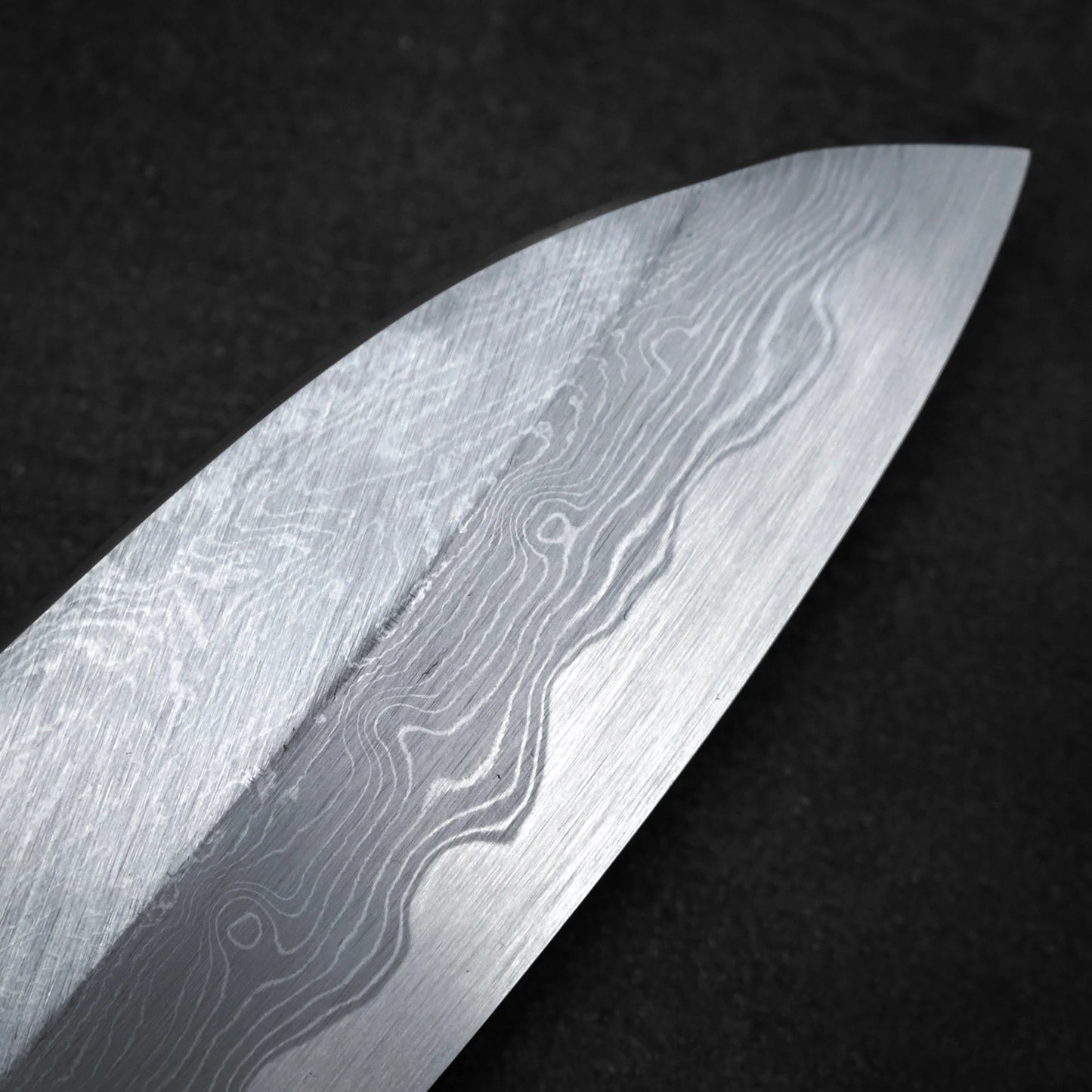 A close-up of the Satoshi Nakagawa Nakagawa aogami#1 damascus santoku 180mm blade, showcasing its striking damascus wave patterns against a dark, textured background.