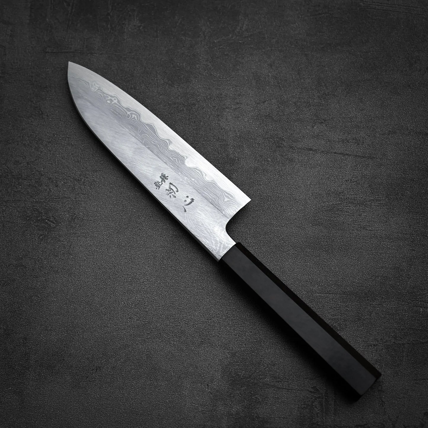The Satoshi Nakagawa Nakagawa aogami#1 damascus santoku 180mm, featuring a damascus finish and black handle, rests on a dark gray textured surface, highlighting its elegant patterned steel blade.