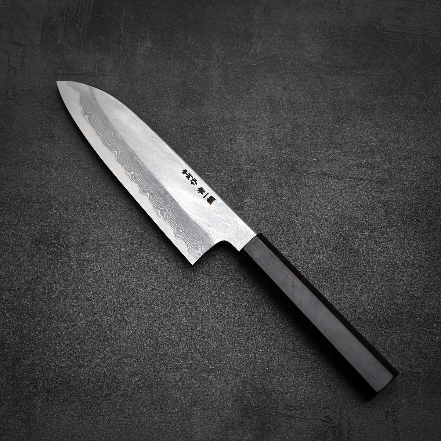 The Satoshi Nakagawa Nakagawa aogami#1 damascus santoku 180mm, featuring a dark wooden handle and elegant patterned steel blade, rests on a textured dark surface.