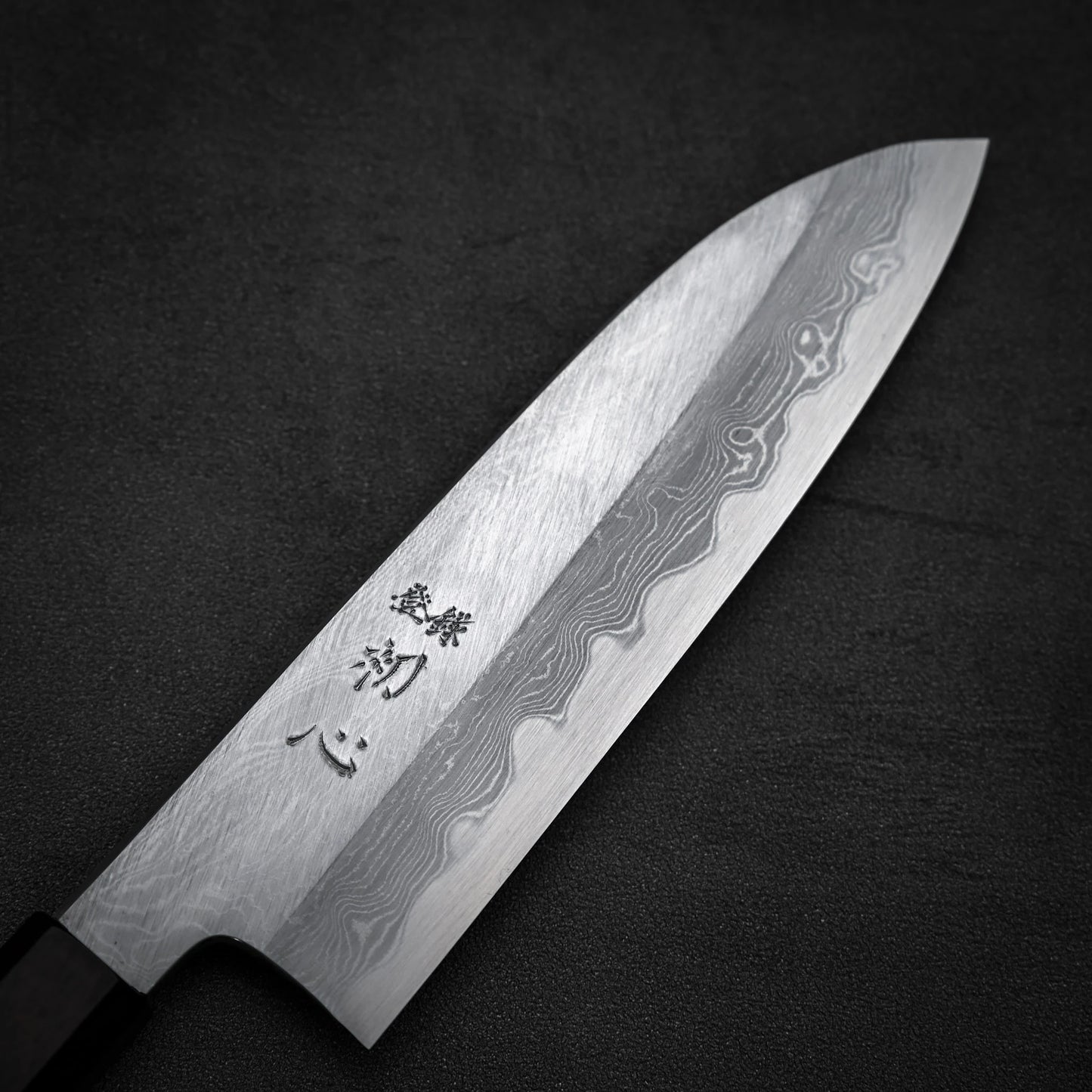A close-up of the Satoshi Nakagawa Nakagawa aogami#1 damascus santoku 180mm knife, featuring a stunning damascus finish and Japanese characters near the handle, set on a dark textured surface.