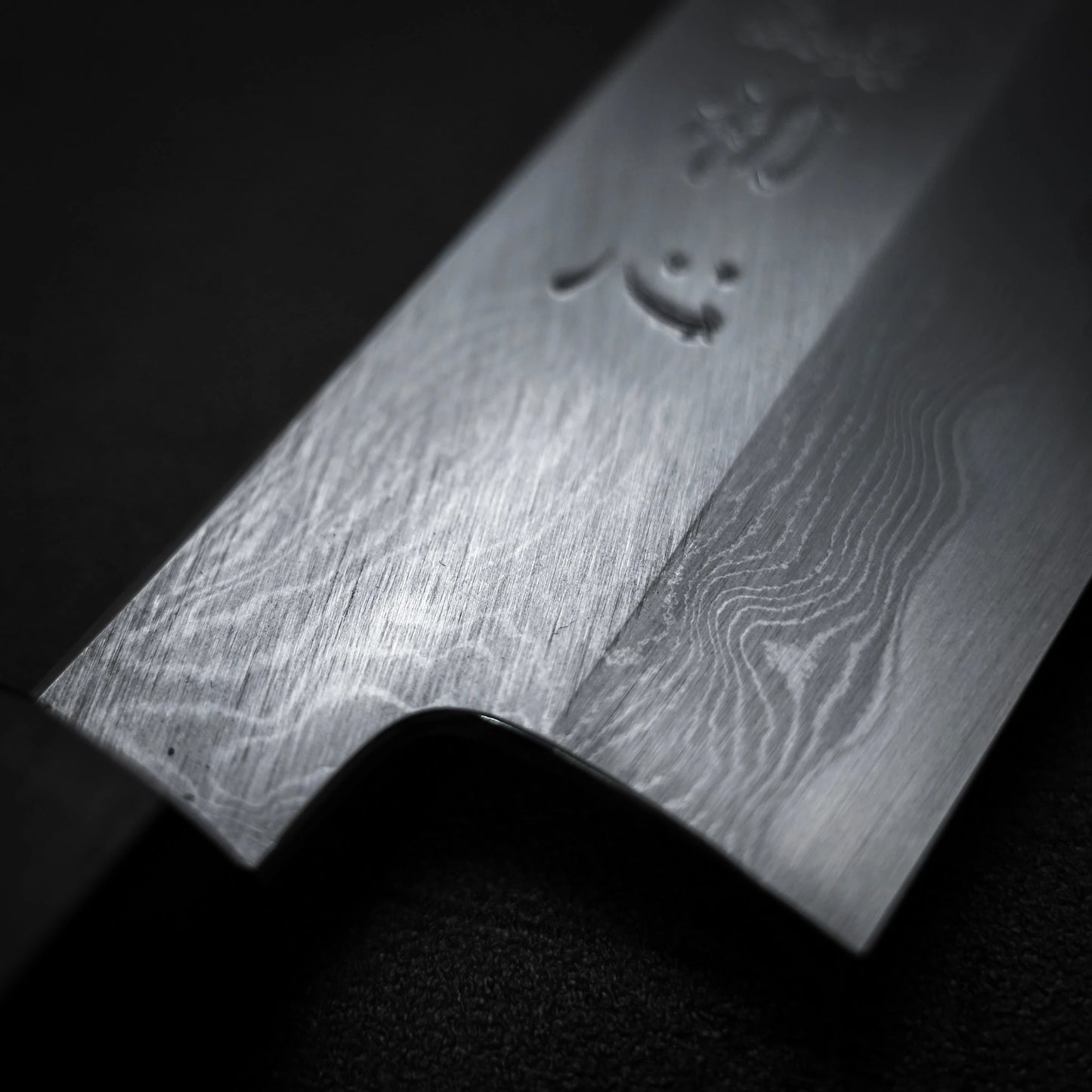 Close-up of the Satoshi Nakagawa Nakagawa aogami#1 damascus santoku 180mm blade, highlighting its detailed damascus patterns and etched markings on a sleek black background.