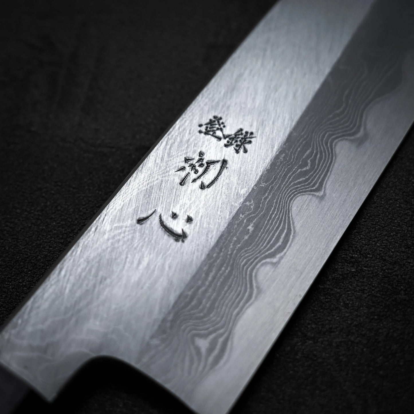 Close-up of the Satoshi Nakagawa Nakagawa aogami#1 damascus santoku 180mm knife blade, featuring a striking damascus finish and engraved Japanese characters, set against a dark background.