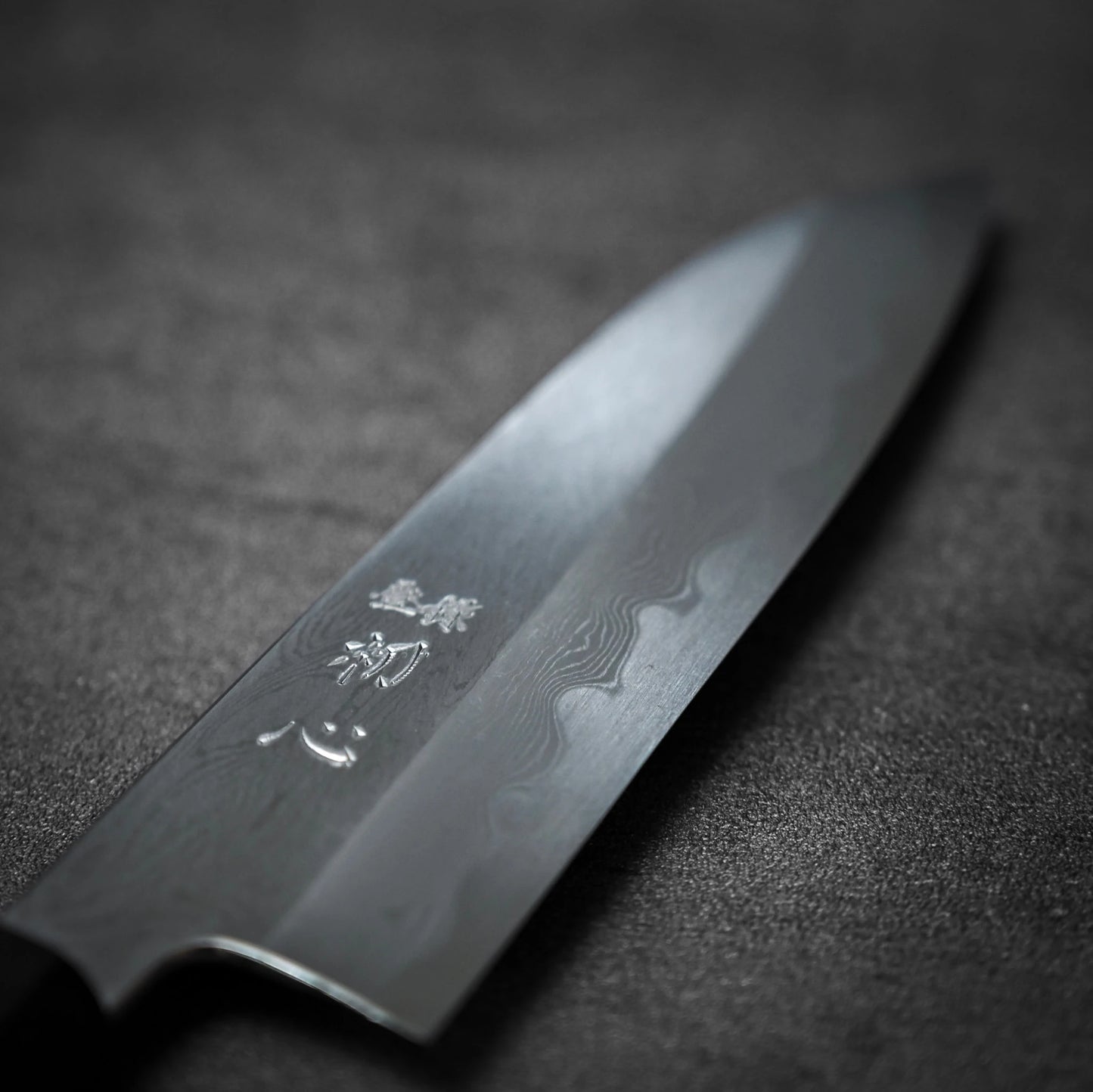 A close-up of the Satoshi Nakagawa Nakagawa aogami#1 damascus santoku 180mm knife, featuring wavy damascus patterns and engraved kanji on the blade, displayed on a dark, textured surface.