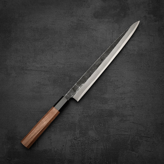 The Hatsukokoro Shirasagi kurouchi aogami#2 yanagiba 270mm, featuring a long, narrow blade and wooden handle, is displayed on a dark, textured surface.