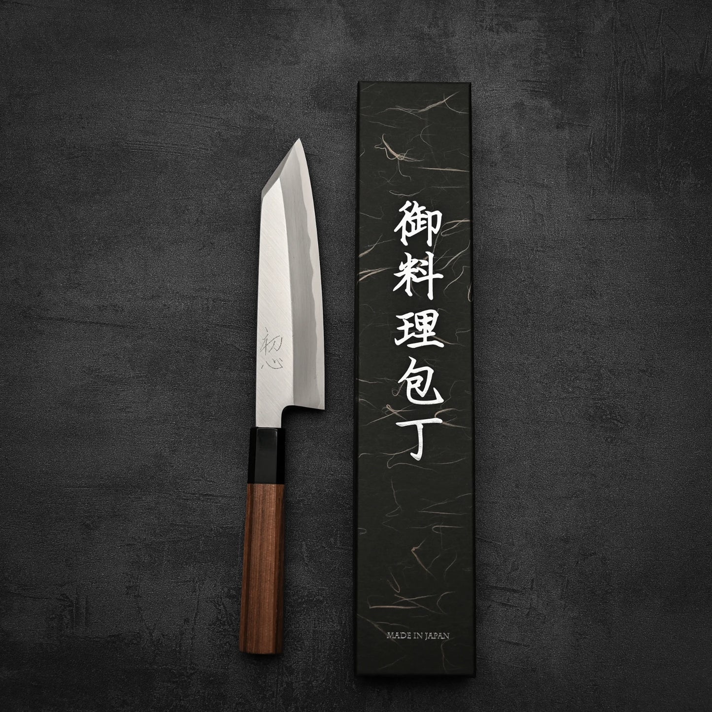 The Hatsukokoro Shirasagi shirogami#2 single bevel kiritsuke gyuto 180mm chefs knife with a wooden handle is placed next to a black box featuring white Japanese characters and MADE IN JAPAN on a dark textured surface.