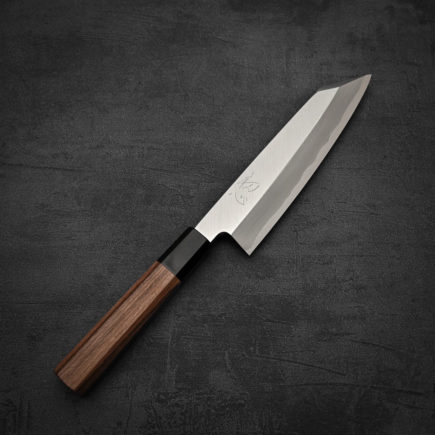 A Hatsukokoro Shirasagi shirogami#2 single bevel kiritsuke gyuto 180mm chefs knife with a wooden handle rests on a dark, textured surface.