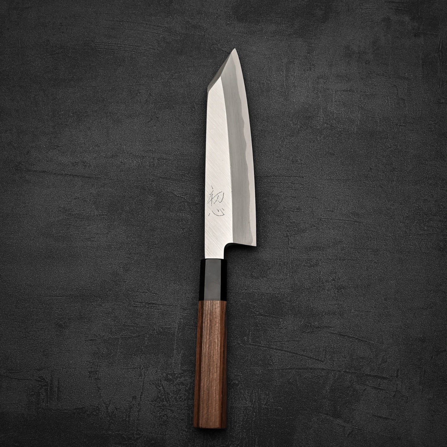 The Hatsukokoro Shirasagi shirogami#2 single bevel kiritsuke gyuto 180mm, a Japanese chefs knife with a wooden handle and shiny angled blade, rests on a dark textured surface.
