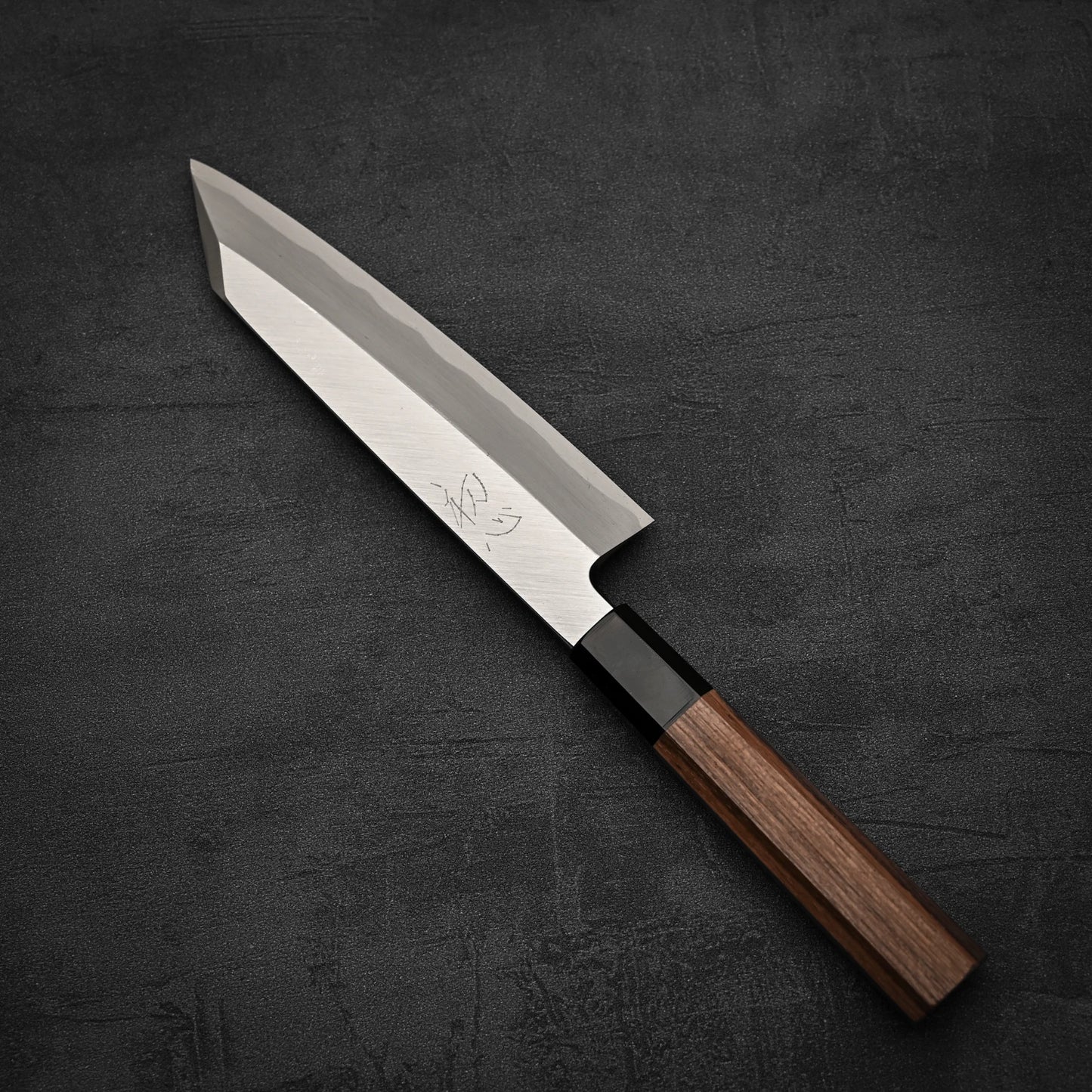 The Hatsukokoro Shirasagi shirogami#2 single bevel kiritsuke gyuto 180mm kitchen knife, featuring a sharp angular blade and a wooden handle, rests on a dark textured surface.