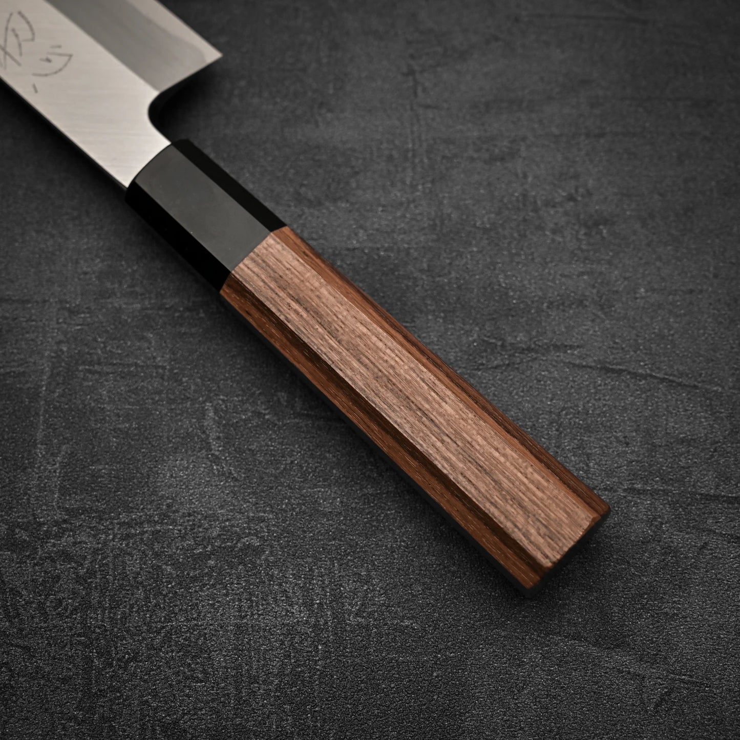 Close-up of the Hatsukokoro Shirasagi shirogami#2 single bevel kiritsuke gyuto 180mm, showing its polished wooden handle and stainless steel blade on a dark textured surface. Only part of the blade and most of the handle are visible.