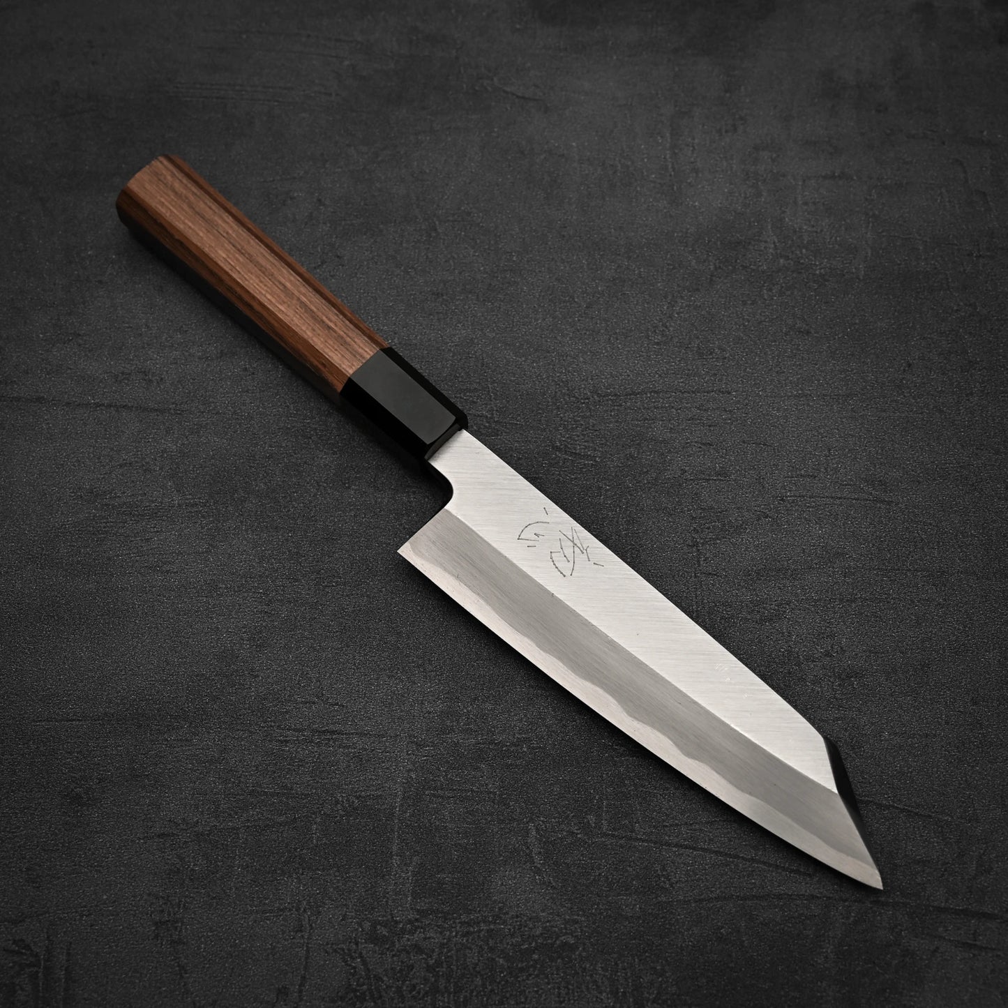 The Hatsukokoro Shirasagi shirogami#2 single bevel kiritsuke gyuto 180mm boasts a wooden handle, a sharp angular blade with subtle engravings, and rests on a dark textured surface.