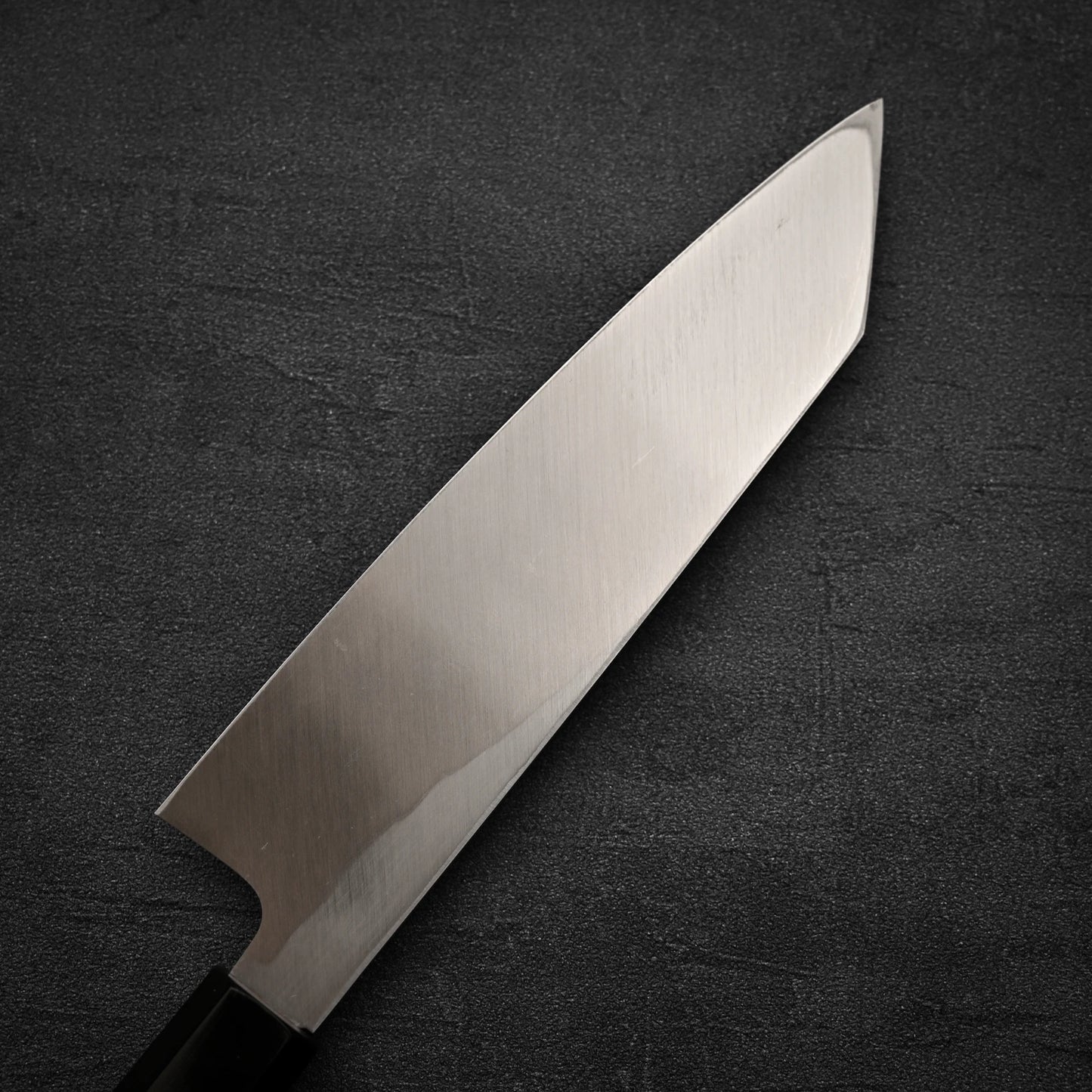 A close-up of the Hatsukokoro Shirasagi shirogami#2 single bevel kiritsuke gyuto 180mm, featuring a sharp blade and black handle, is displayed on a dark textured surface.