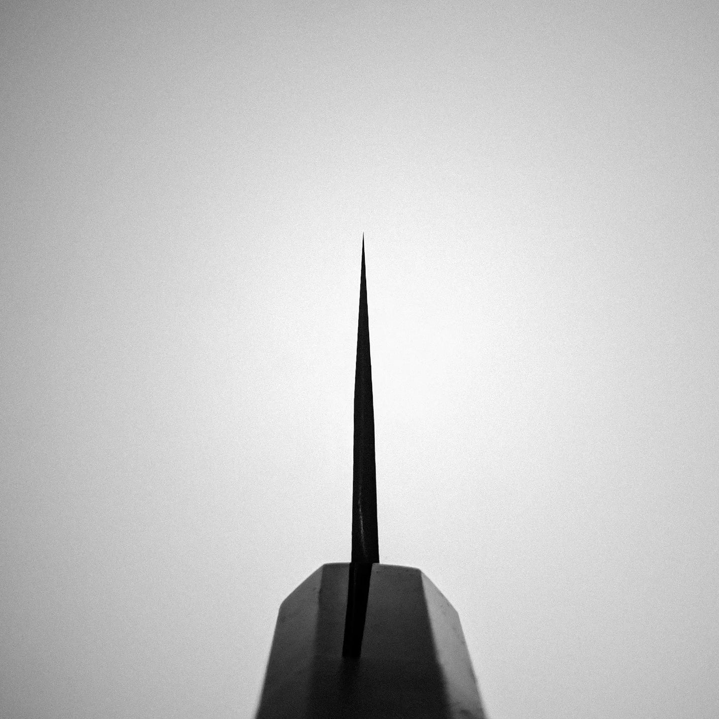 A close-up, upward-facing view of the tip of a Yoshikane Hatsukokoro x Yoshikane nashiji shirogami#2 kiritsuke gyuto 210mm Japanese knife against a plain, light grey background, centered in the frame.